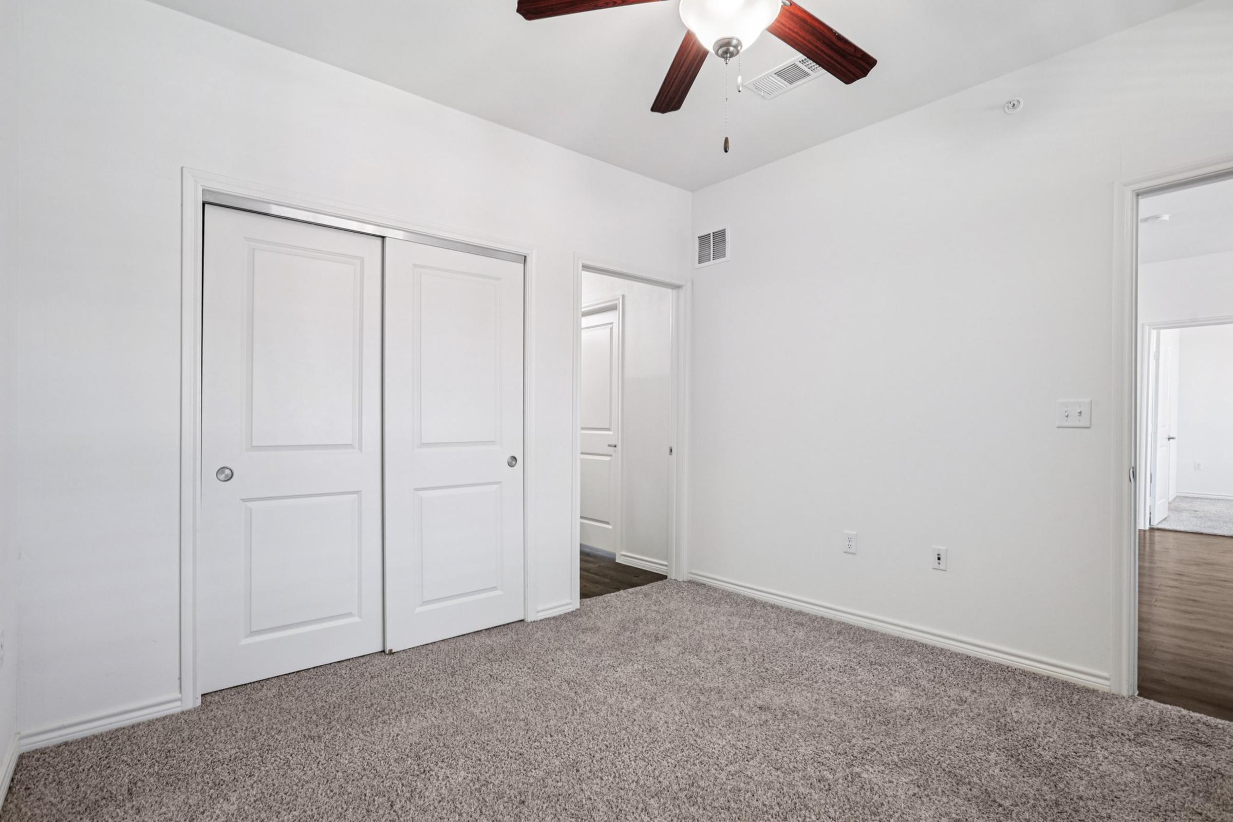 A well-lit, empty room featuring carpeted flooring, a ceiling fan, and two white double doors leading to a closet. The walls are painted a light color, and there is another doorway visible in the background, suggesting access to additional spaces.