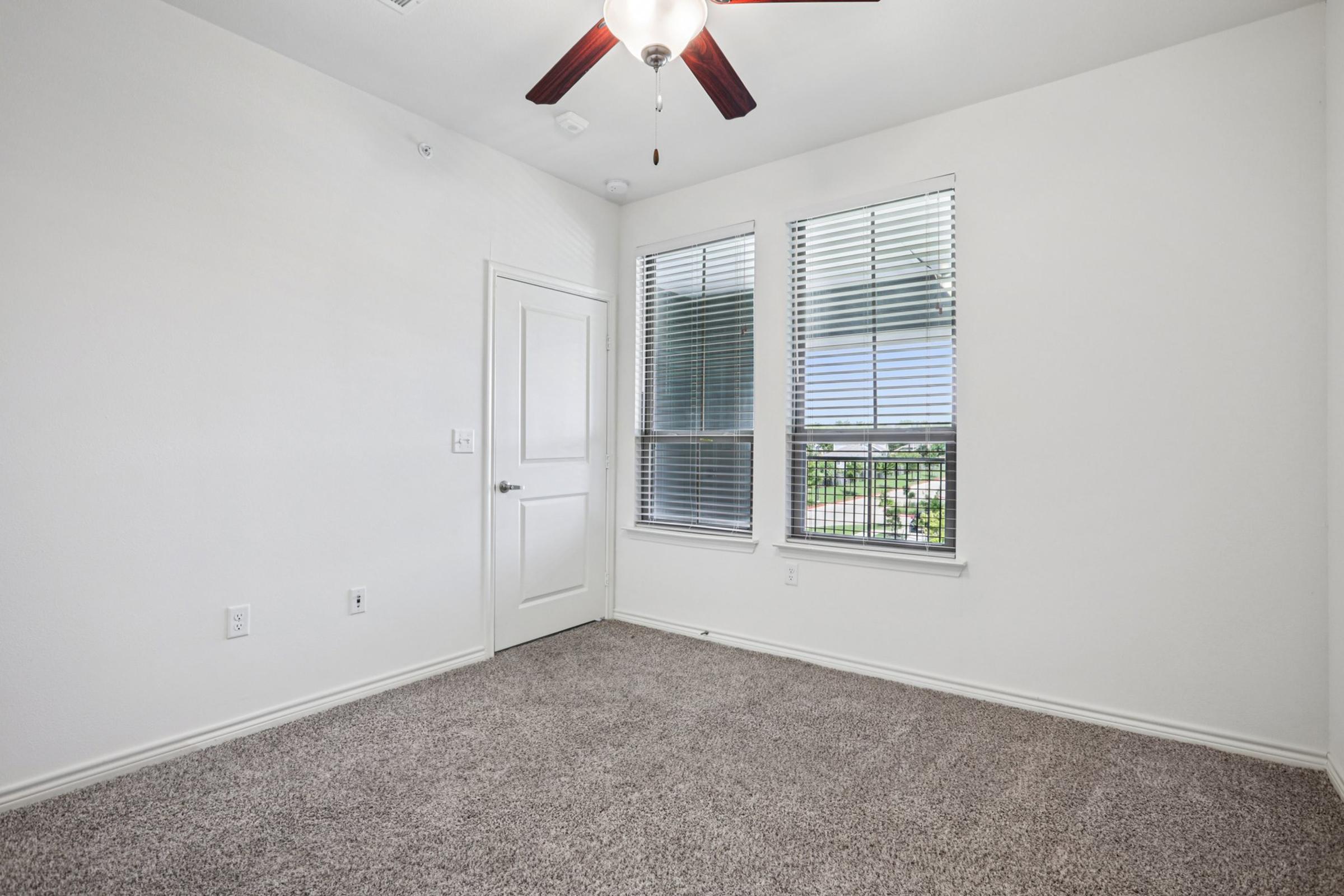 A bright and empty room with light-colored walls and carpet. It features two windows with white blinds, allowing natural light to enter. A ceiling fan with wooden blades hangs above, and a closed door leads to another space. The room appears freshly painted and spacious, suitable for various purposes.