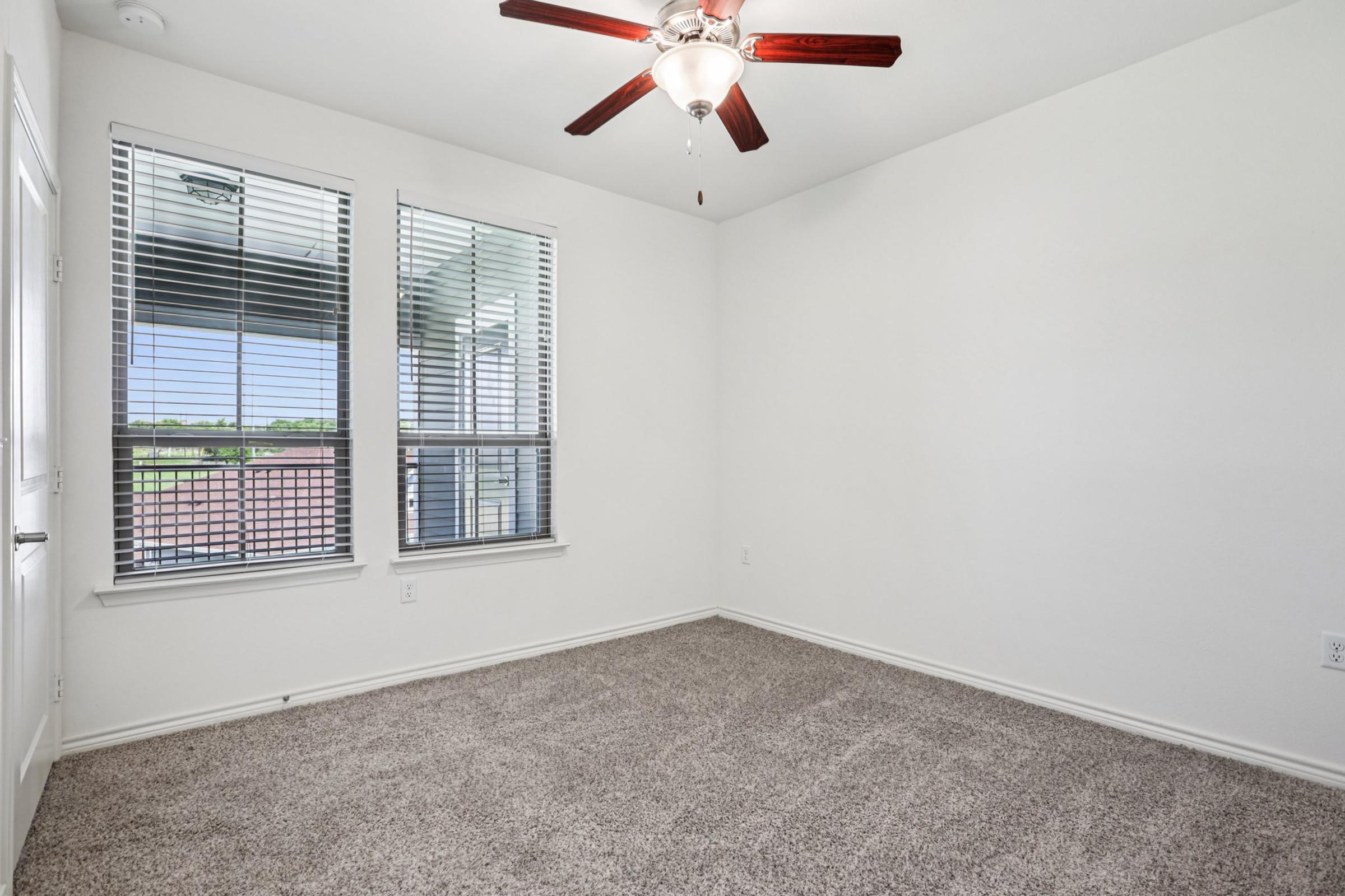 A well-lit room with beige carpet, featuring a ceiling fan with wooden blades. There are two windows with blinds, allowing natural light to fill the space. The walls are painted white, creating a clean and airy atmosphere. The room is empty, suggesting it can be used for various purposes, like a bedroom or office.