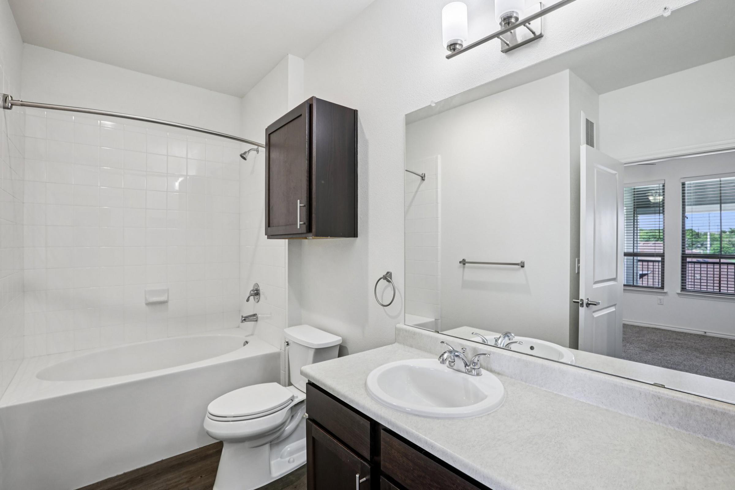 A modern bathroom featuring a white bathtub with a shower, a dark wood vanity with a sink, and a large mirror. The area is well-lit with natural light from a window. Neutral-colored walls and tiled flooring create a clean and contemporary look.