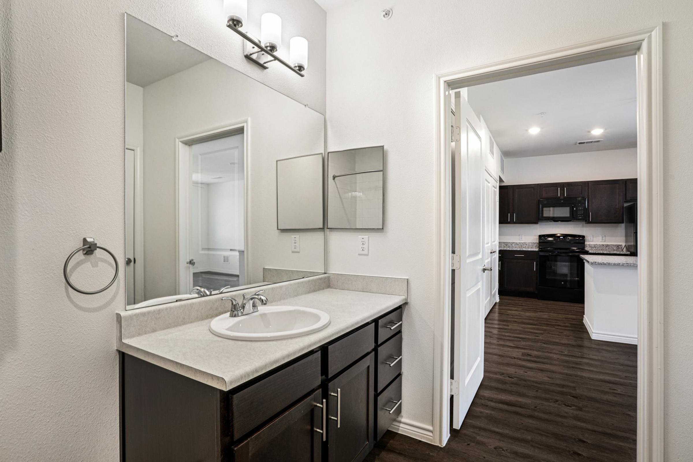 A modern bathroom featuring a vanity with a sink and mirror, dark wood cabinetry, and neutral-colored walls. A doorway leads to a kitchen area with dark cabinets and a black stove visible in the background. The flooring is dark, adding contrast to the lighter walls.
