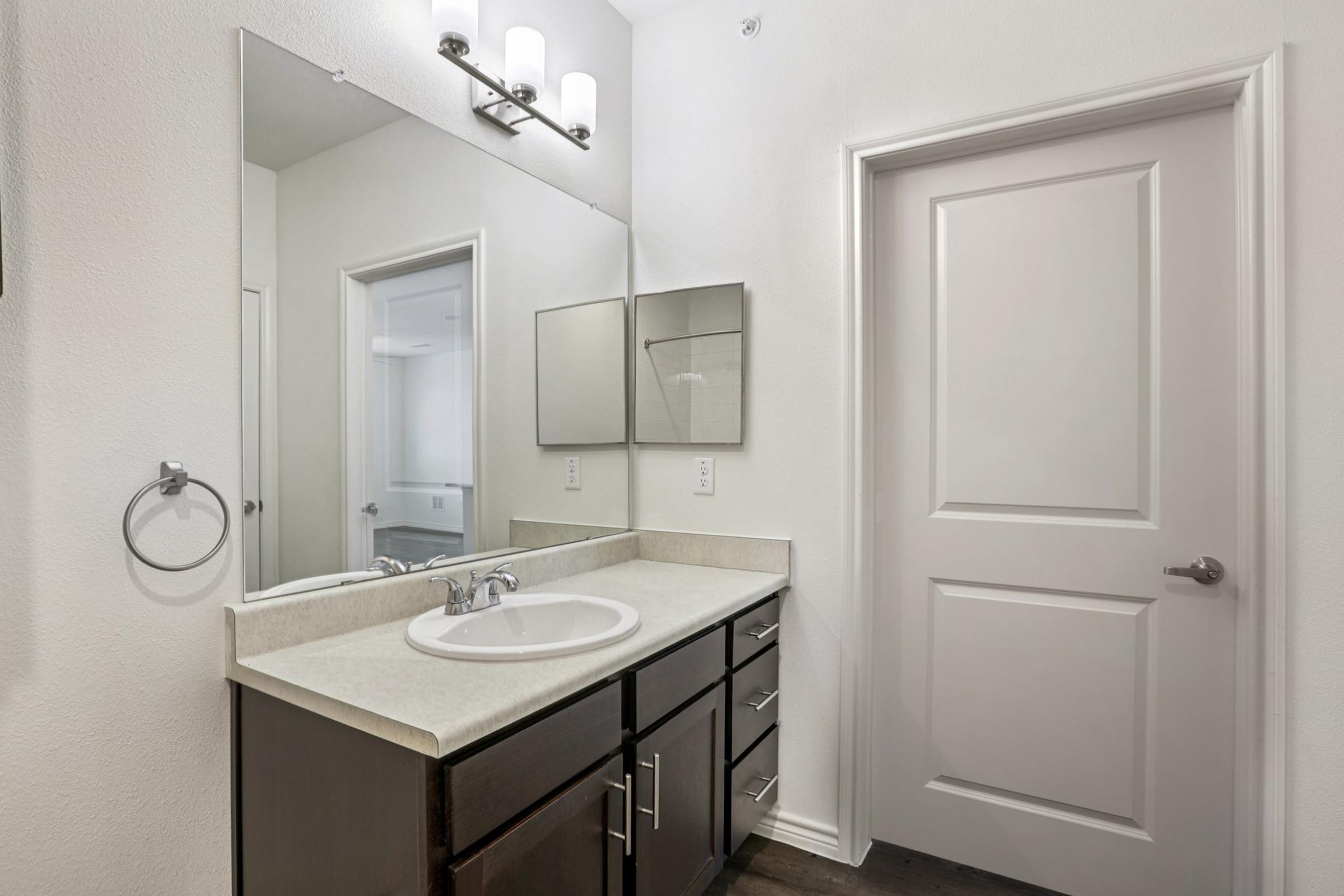 A modern bathroom with a white countertop and dark wood cabinet. The wall features a large mirror above the sink, along with a light fixture. A closed door is visible on the side, along with a towel ring. The walls are painted in a light color, creating a clean, bright atmosphere.