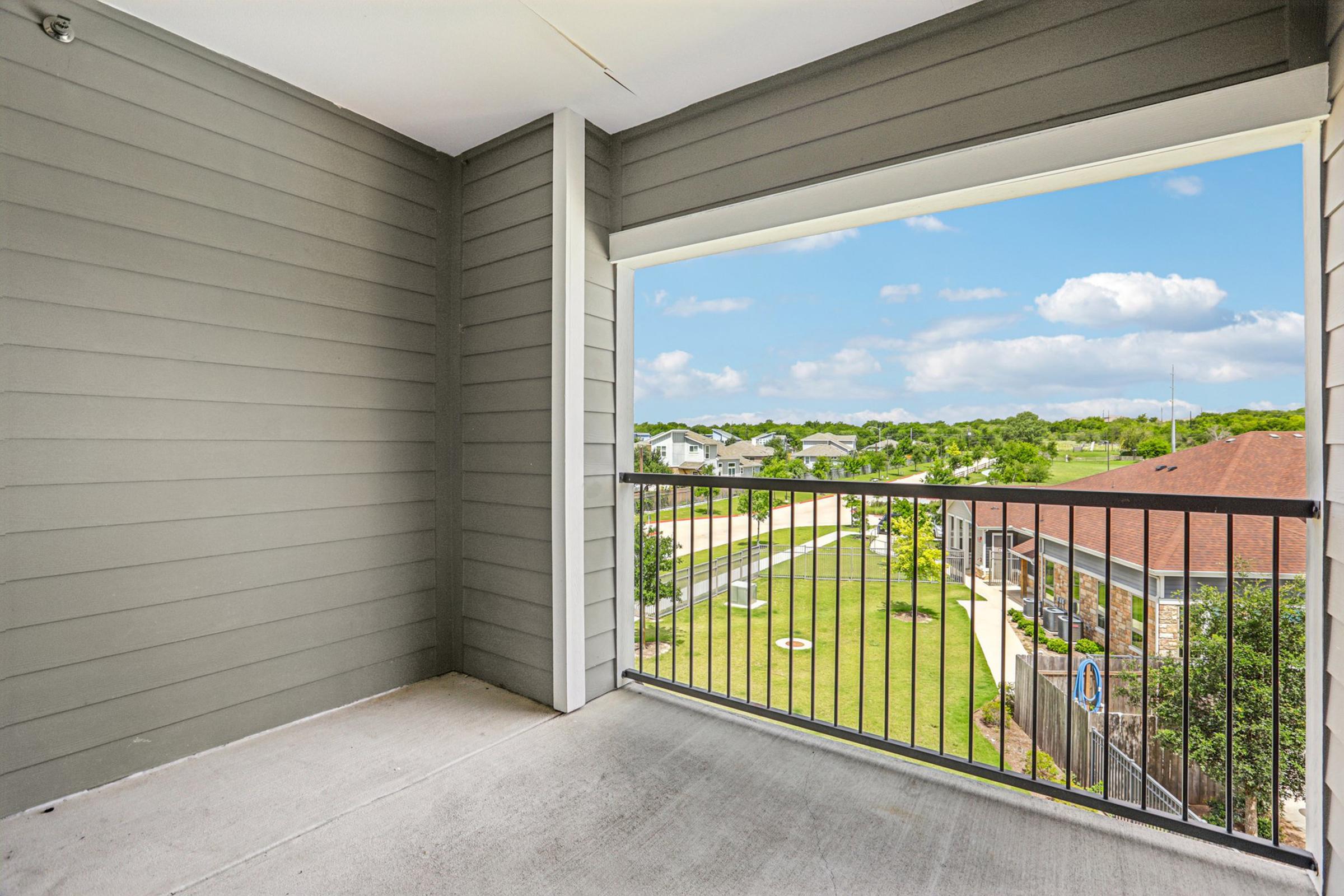 View from a second-floor balcony showcasing a spacious outdoor area with a gray exterior wall. The balcony features a black metal railing and overlooks a green lawn and nearby buildings under a blue sky with scattered clouds.