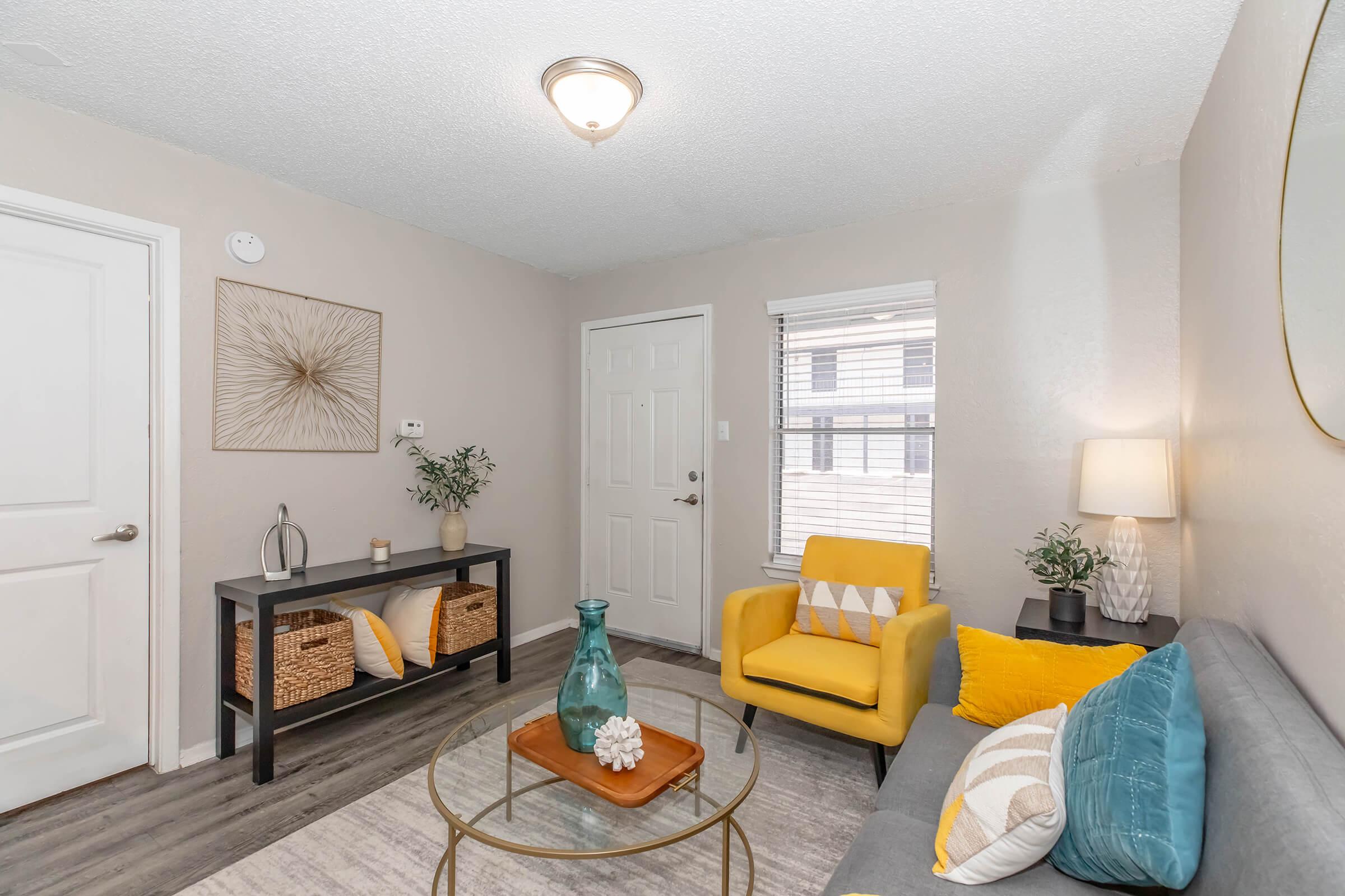 Cozy living room featuring a gray sofa with decorative cushions, a yellow chair, a small coffee table with a vase, and a console table with baskets. The walls are light-colored, and there’s a door leading outside. Bright natural light enters through the window. A modern light fixture is visible on the ceiling.