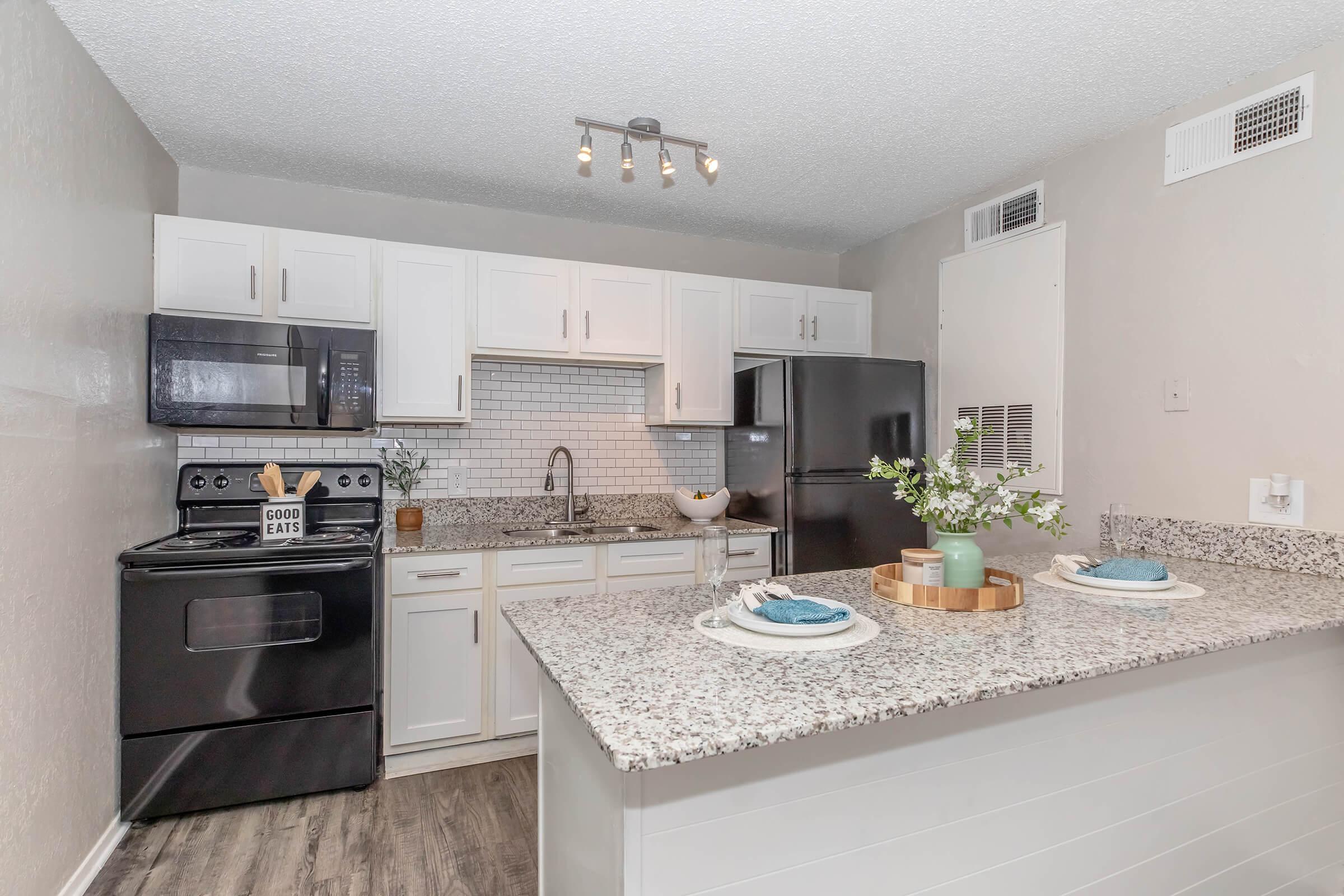 A modern kitchen featuring white cabinetry, a black stove, and a stainless steel refrigerator. The countertop is made of gray speckled granite, and there are decorative items including a plant and a wood serving tray with plates. Overhead lighting adds brightness to the space.