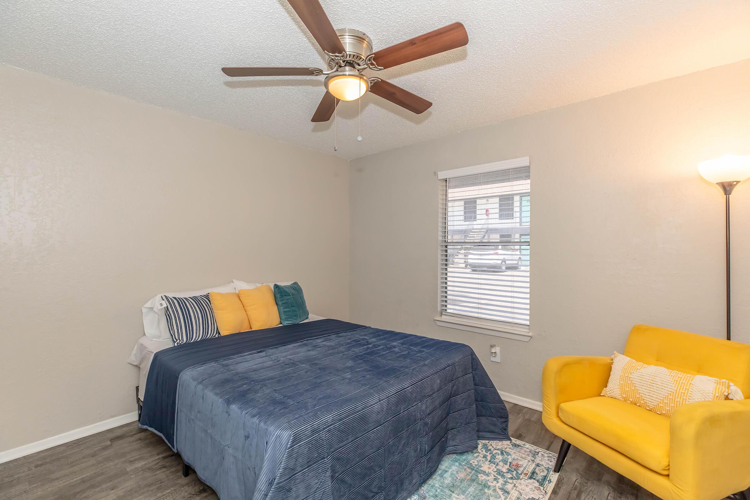 A cozy bedroom featuring a bed with blue bedding and colorful pillows, a ceiling fan, a yellow armchair, and a window with blinds. The room has light gray walls and a decorative area rug on the floor.