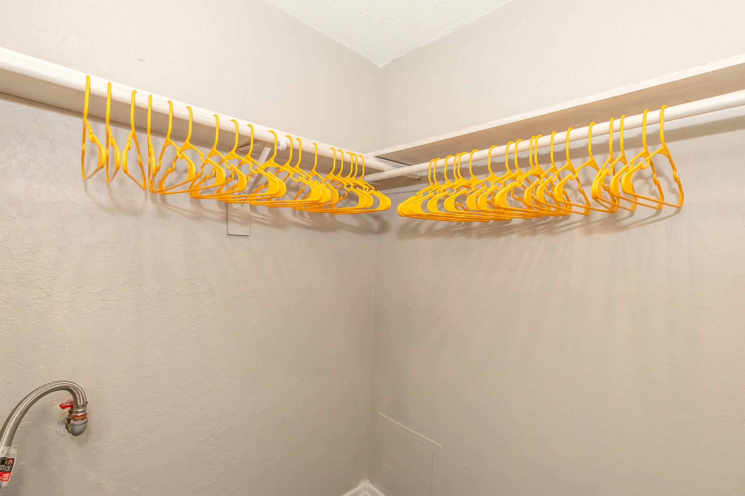 A close-up view of a neatly organized closet with a white rod holding several yellow plastic hangers, all arranged in a row against a plain beige wall. A silver pipe is visible on the left side of the image.