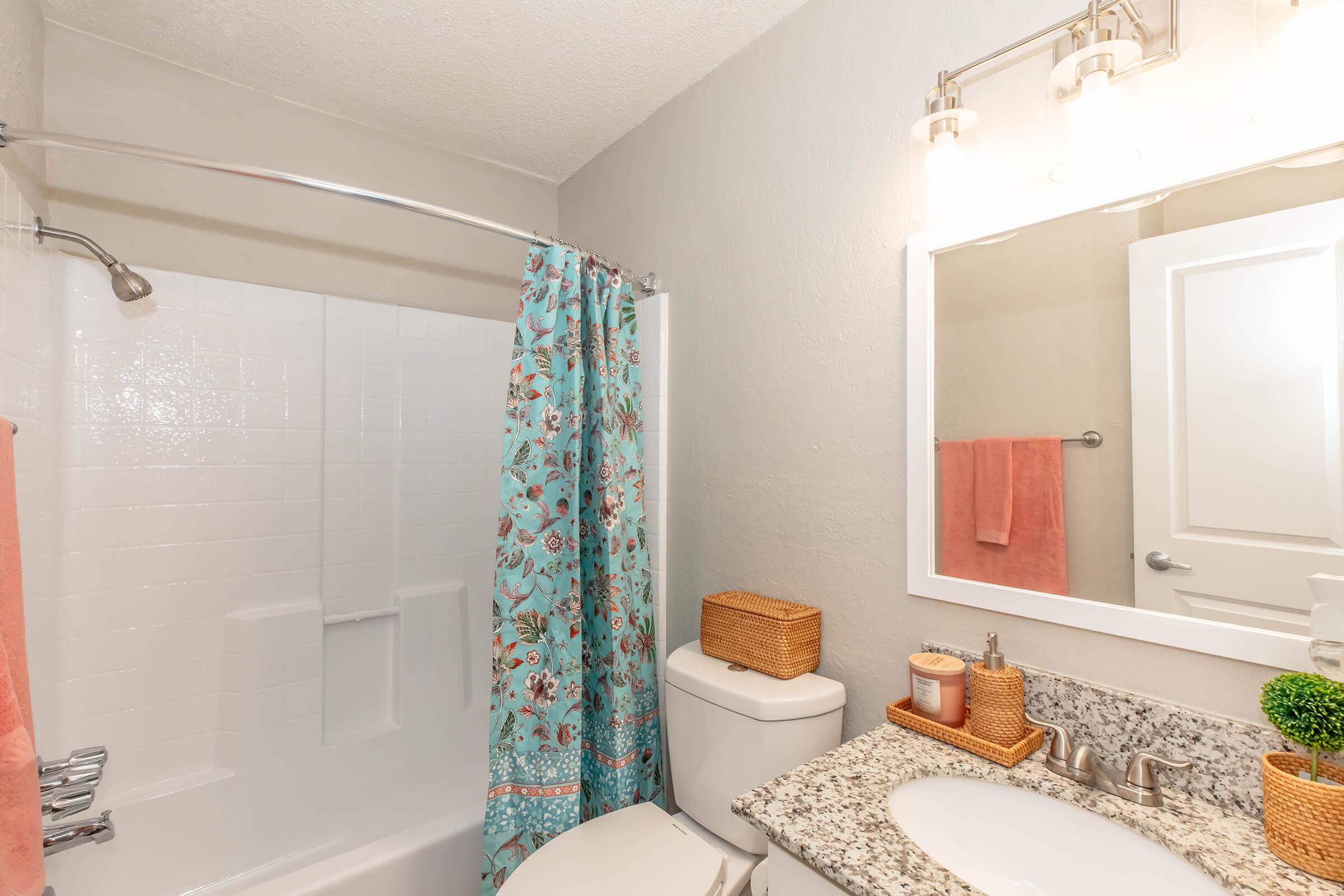 A bathroom featuring a white bathtub with a floral patterned shower curtain, a granite countertop with a sink, and a mirror above. The walls are painted light gray, and there are decorative elements like a pink towel and wicker basket. Soft lighting is provided by a fixture above the mirror.