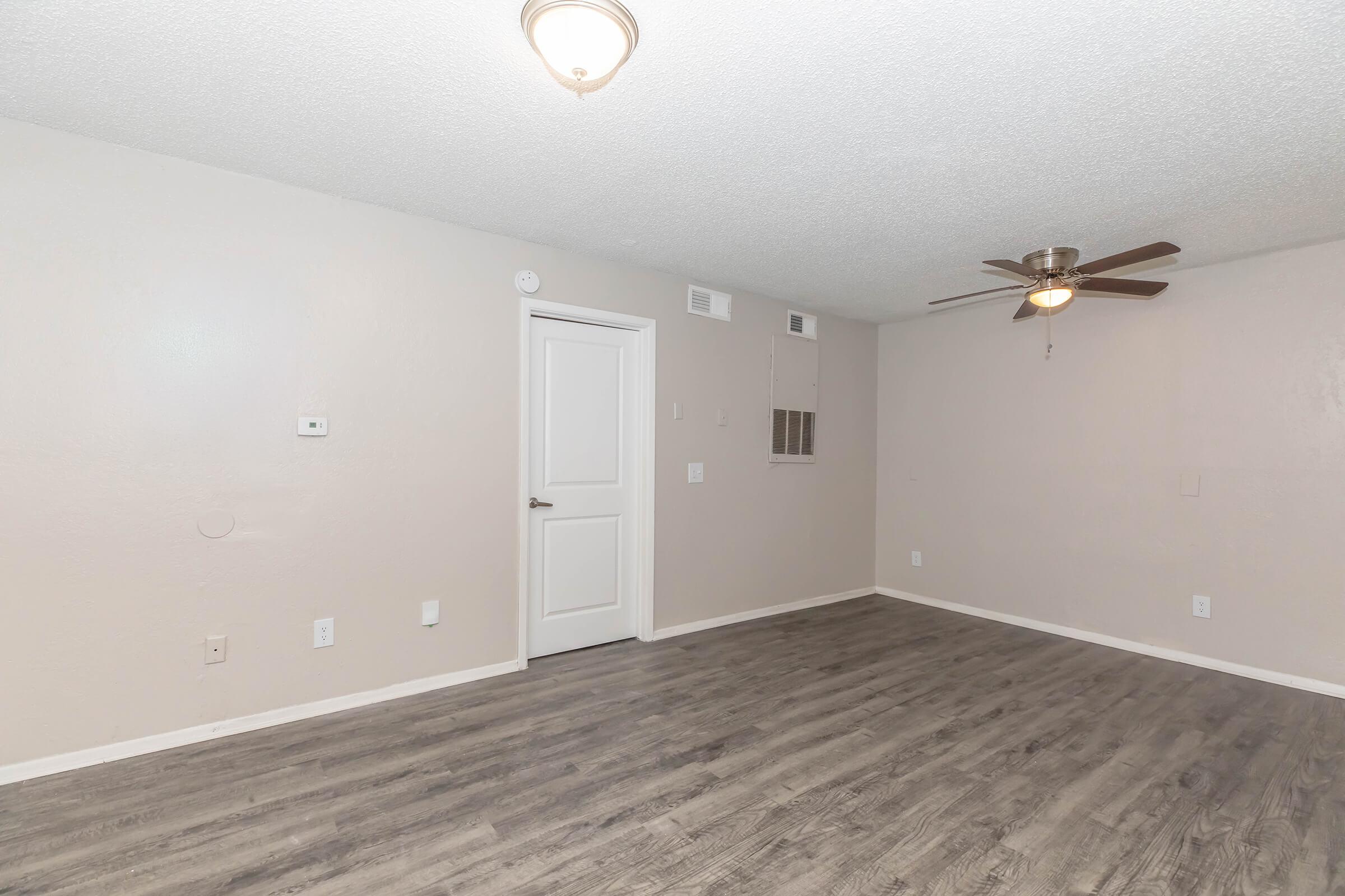 A spacious, empty room with light-colored walls and wood laminate flooring. A ceiling fan is mounted in the center, and there is a door on one side. The room includes a small air vent and light fixtures, creating a bright and airy atmosphere.