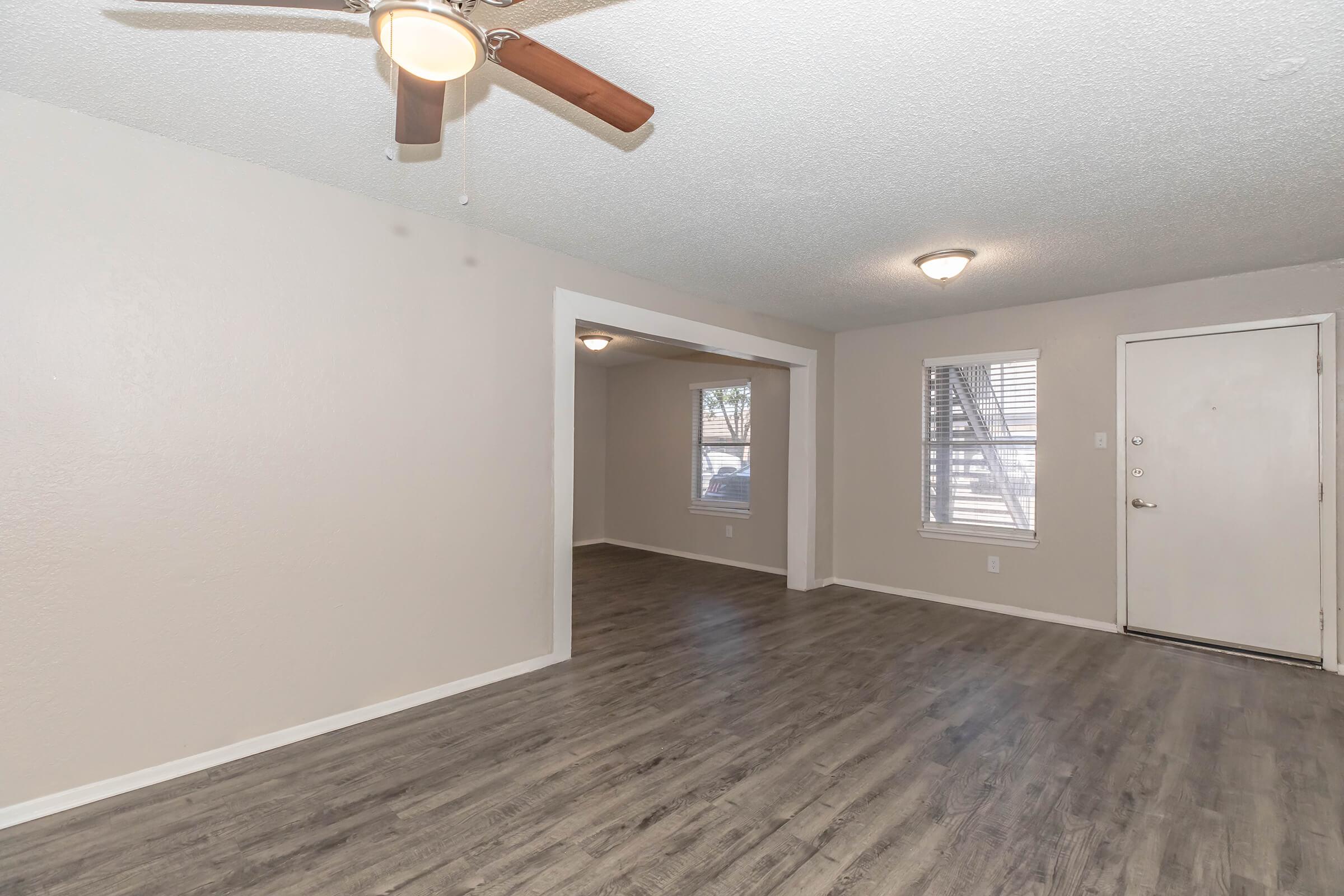 Interior view of a living room featuring light-colored walls, a ceiling fan, and laminate flooring. There is a doorway leading to another room, and a front door with a window nearby. Natural light comes through the windows, creating a bright and open atmosphere.