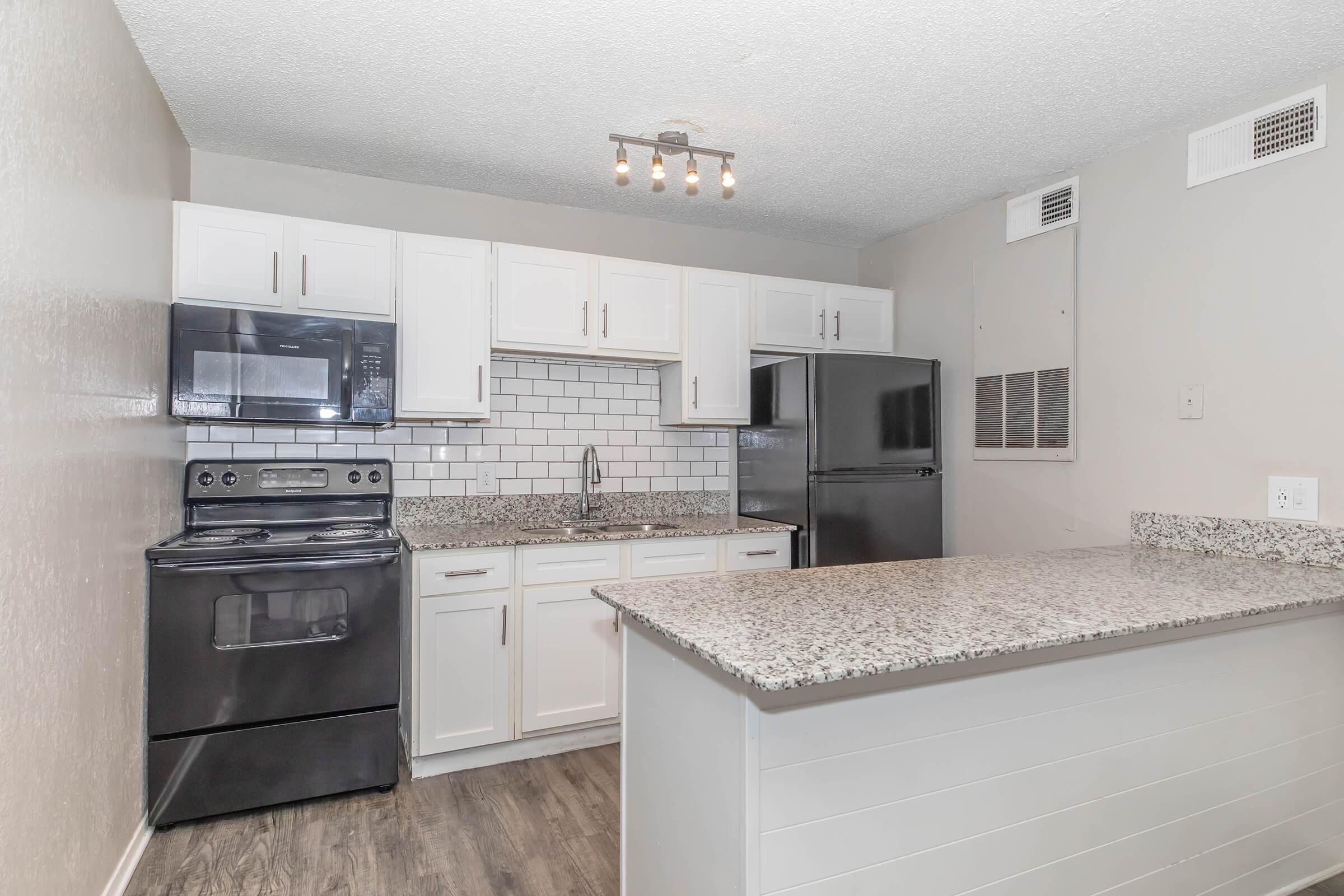 Modern kitchen featuring white cabinets, a black stove and microwave, a granite countertop, and stainless steel appliances. The backsplash is white subway tile, and there is a spacious island with seating. The walls are painted light gray, and the room is well-lit with overhead lighting.