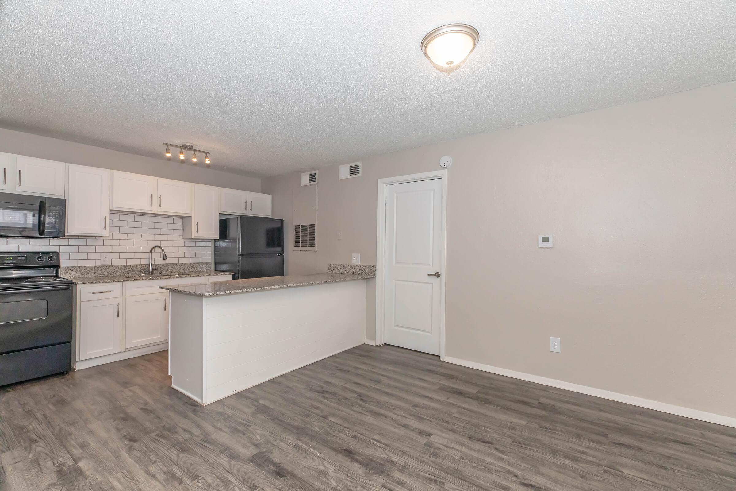 A modern kitchen with white cabinets, a granite countertop, and stainless steel appliances. The space features a dark floor, pendant lighting, and a doorway leading to another room. It has a clean, open layout, ideal for cooking and entertaining.