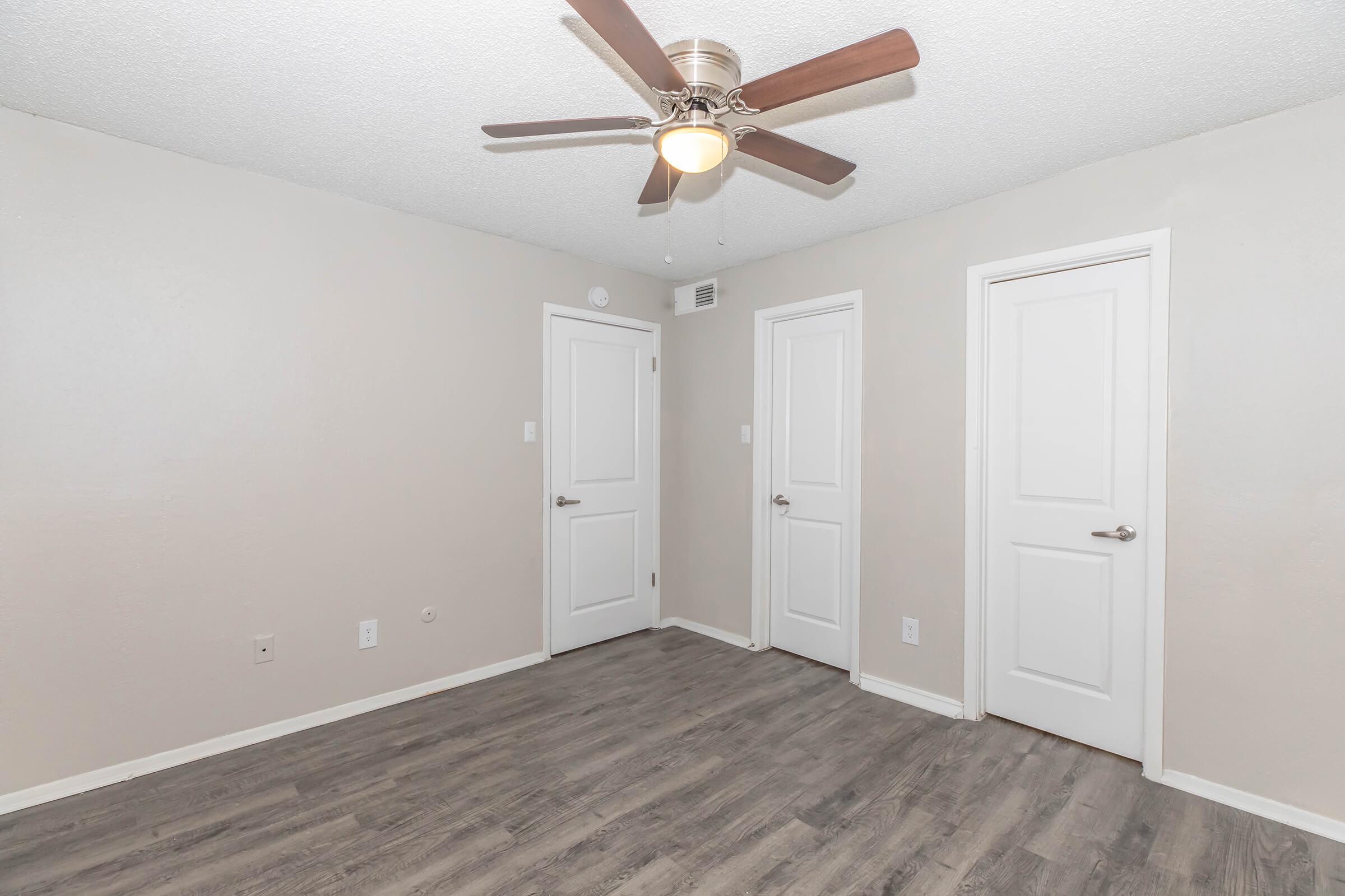 A light-colored room with textured walls, featuring a ceiling fan with wooden blades. There are two closed white doors on the right, and a smooth wooden floor. The space is empty and well-lit, creating a clean and modern atmosphere.