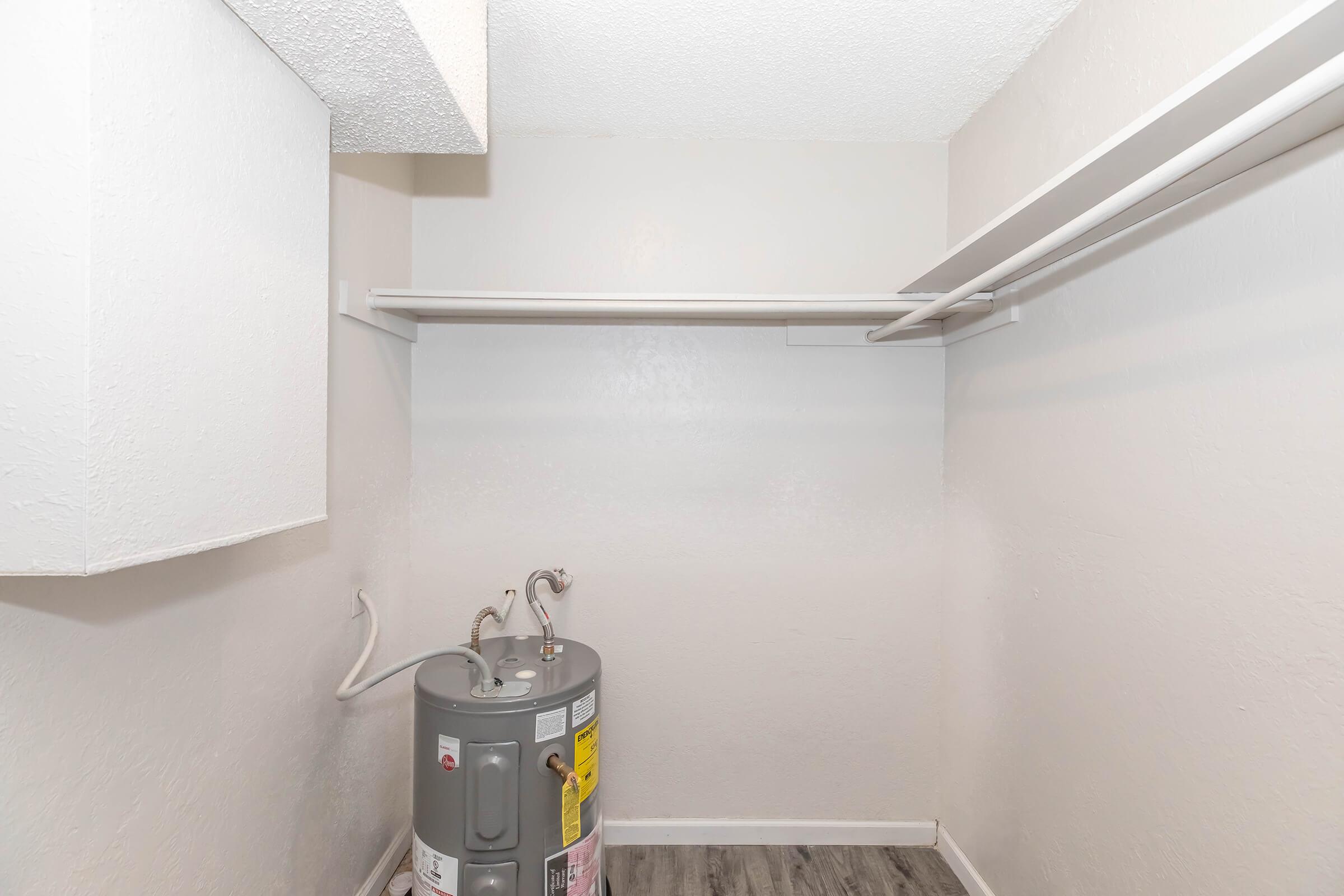 A small, empty utility closet featuring a water heater on the floor and a single clothes rod mounted on the wall. The walls are painted in a light color, and the flooring appears to be laminate. The space is well-lit and uncluttered, suitable for storage or utility purposes.