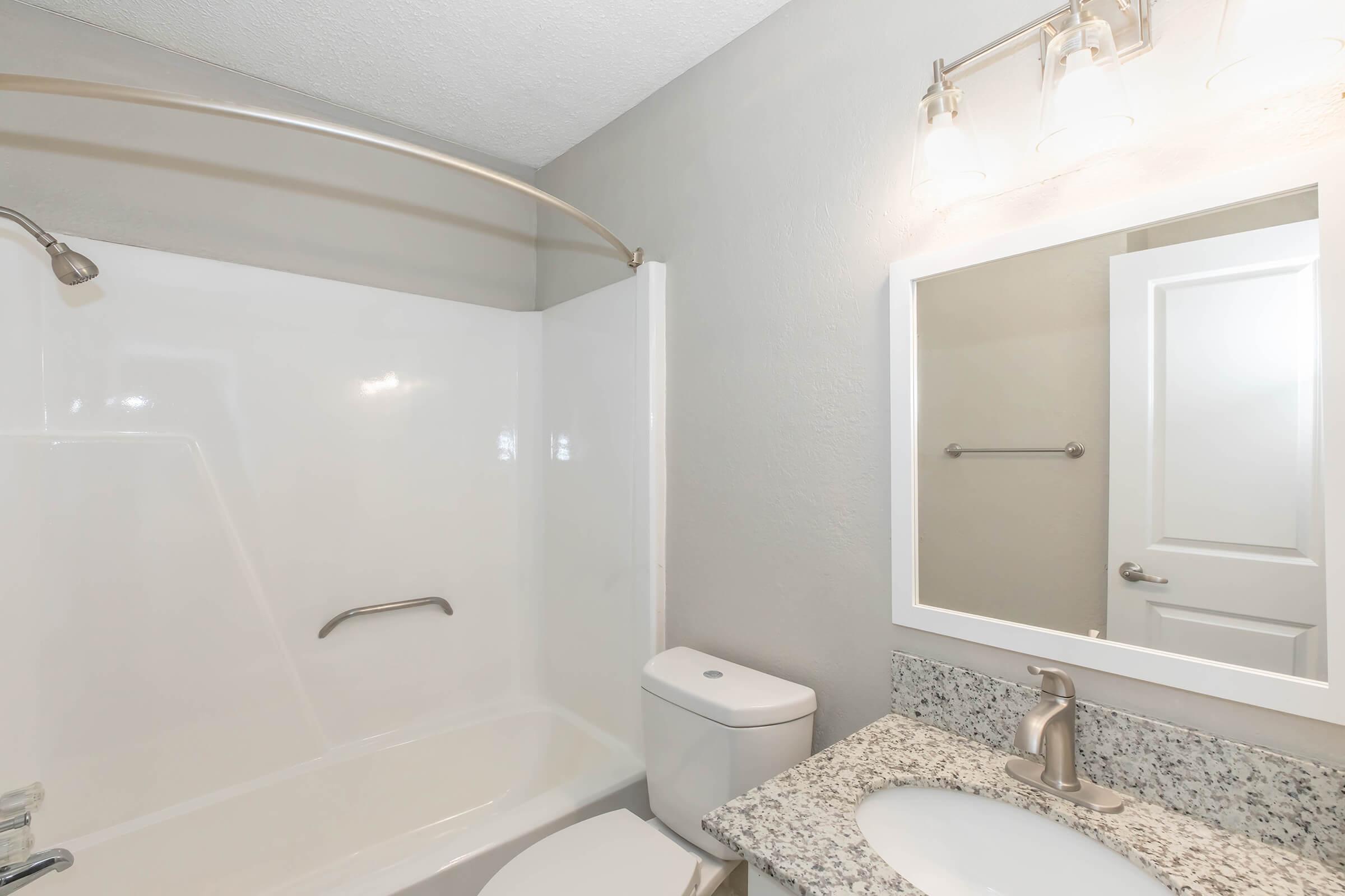 A clean and modern bathroom featuring a shower tub combo with a curved shower rod, a white toilet, a granite countertop sink with a silver faucet, a mirror with three light fixtures above, and a gray painted wall. The overall atmosphere is bright and inviting.