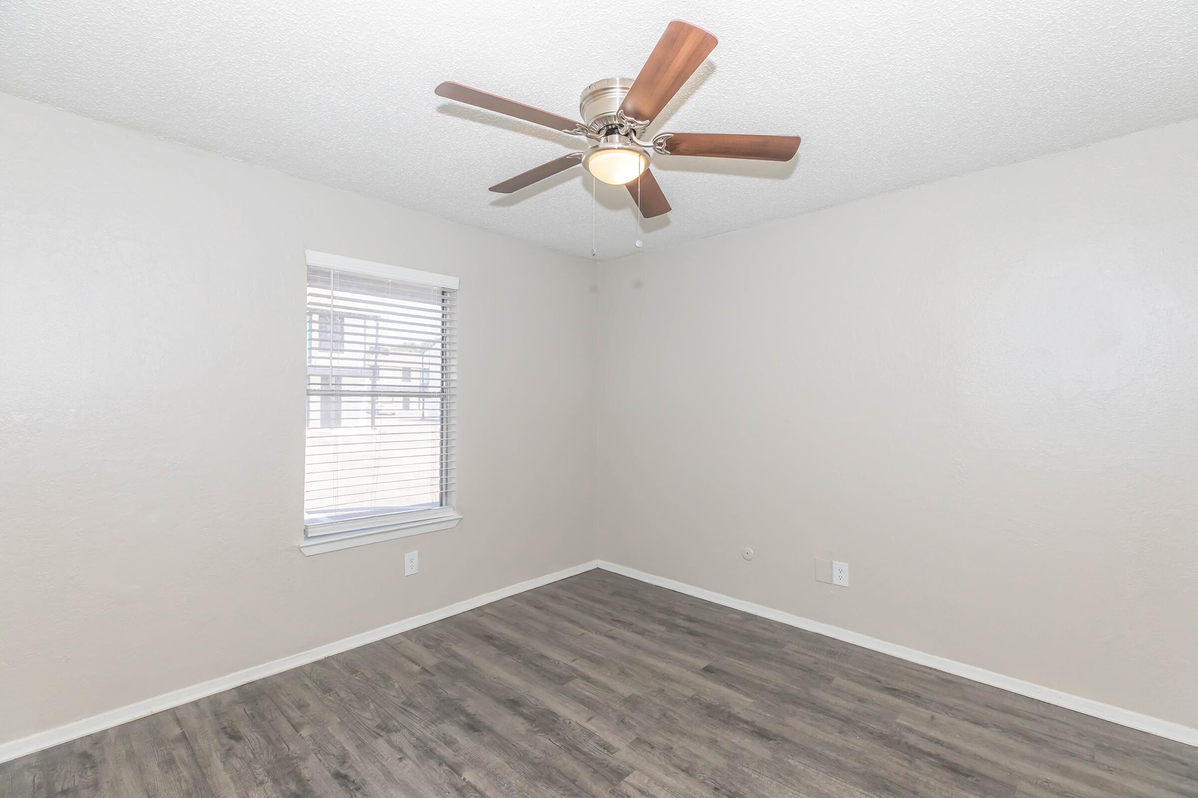 A well-lit, empty room featuring a ceiling fan with wooden blades. The walls are painted a light color, and there is a single window with blinds allowing natural light to enter. The floor is covered with a wood-like laminate flooring, providing a warm and inviting atmosphere.