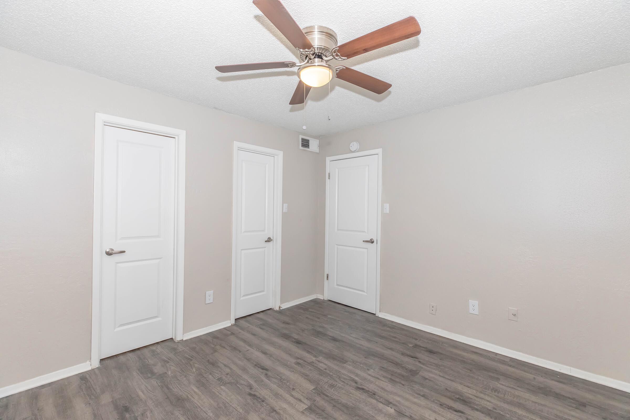 A neutral-colored room with a ceiling fan, featuring three white doors on the walls and light wood laminate flooring. The space is well-lit and appears to be empty, giving it a clean and spacious look.