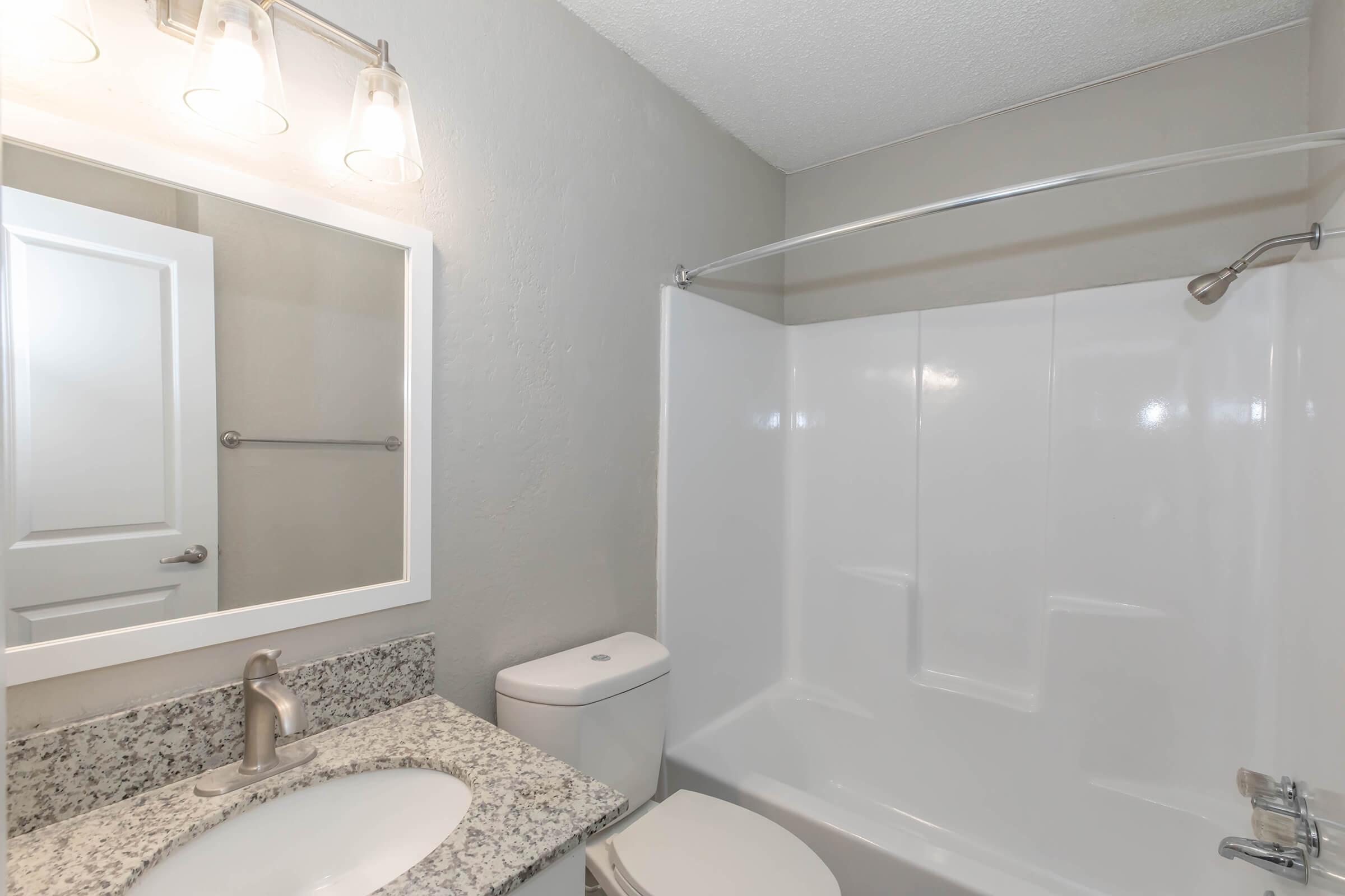 Bright and modern bathroom featuring a white bathtub/shower combination, a compact toilet, and a granite countertop sink area. The walls are painted a light gray, with a square mirror above the sink and stylish lighting fixtures. The clean and simple design offers a contemporary feel.