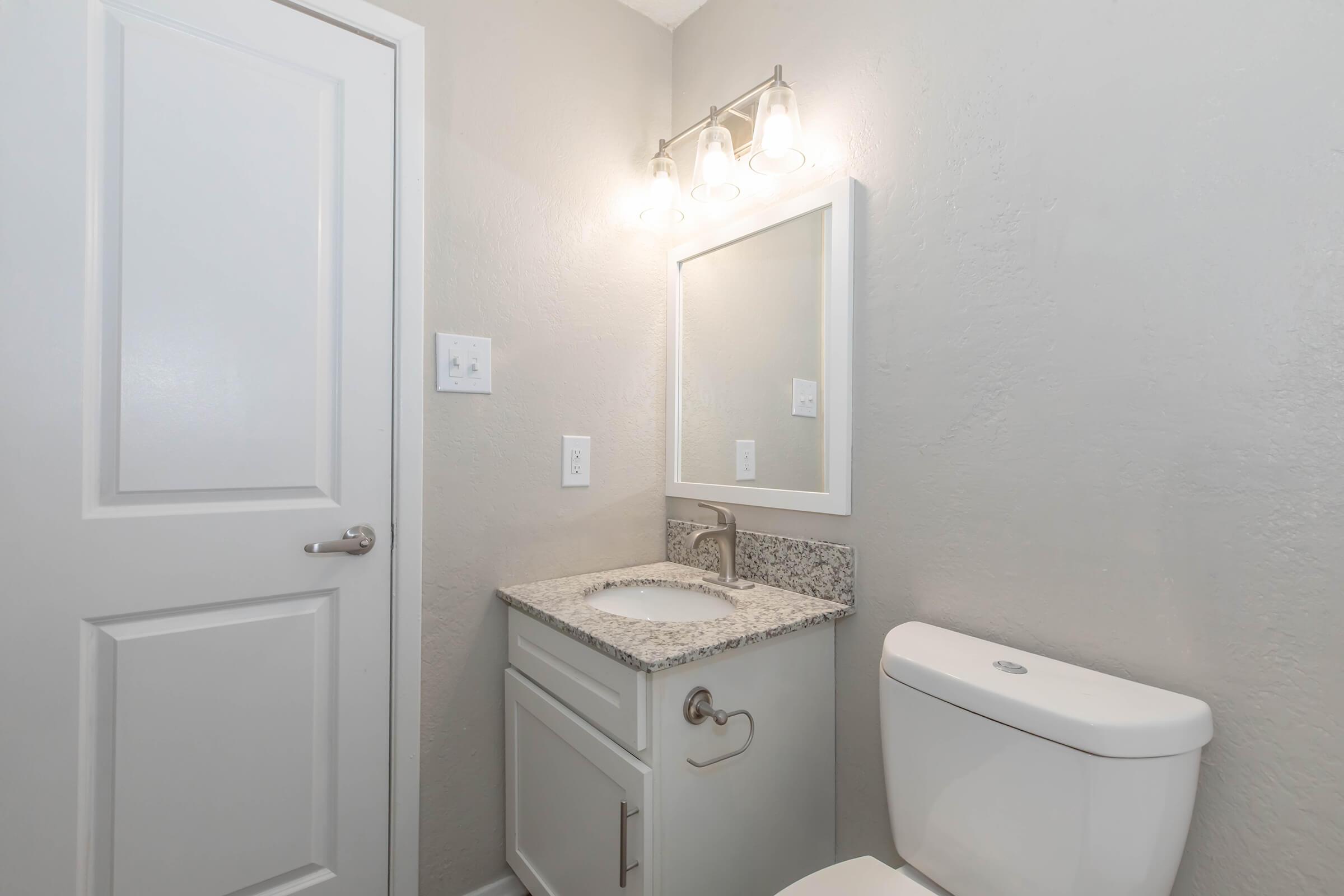 A modern bathroom featuring a white toilet, a gray granite countertop with a sink, and a wall-mounted mirror. The walls are painted light gray, and there is a stylish light fixture above the mirror. A white door is visible on the left side of the image.