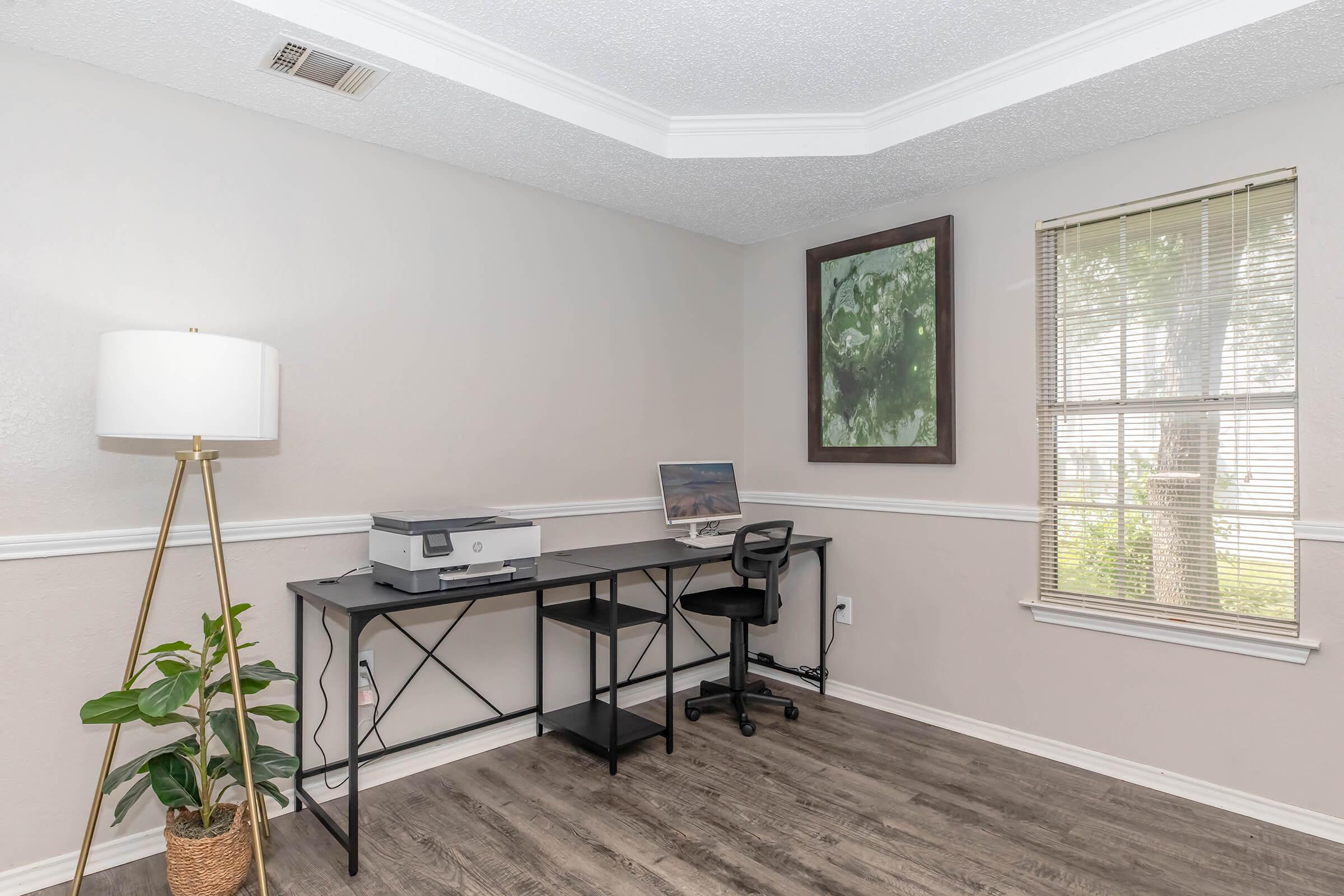 A minimalist home office featuring a black desk with a printer, a black chair, and a decorative plant. A framed nature picture hangs on the wall, and a window provides natural light. The room has light-colored walls and wooden flooring, creating a spacious, inviting atmosphere.