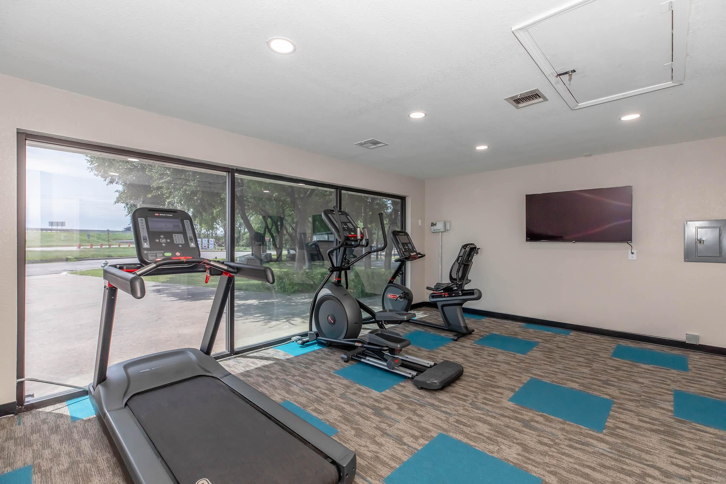 A modern gym featuring a treadmill, elliptical machine, and stationary bike, with large windows offering natural light and views of the outside. The floor has a patterned blue carpet, and there is a wall-mounted television for entertainment.