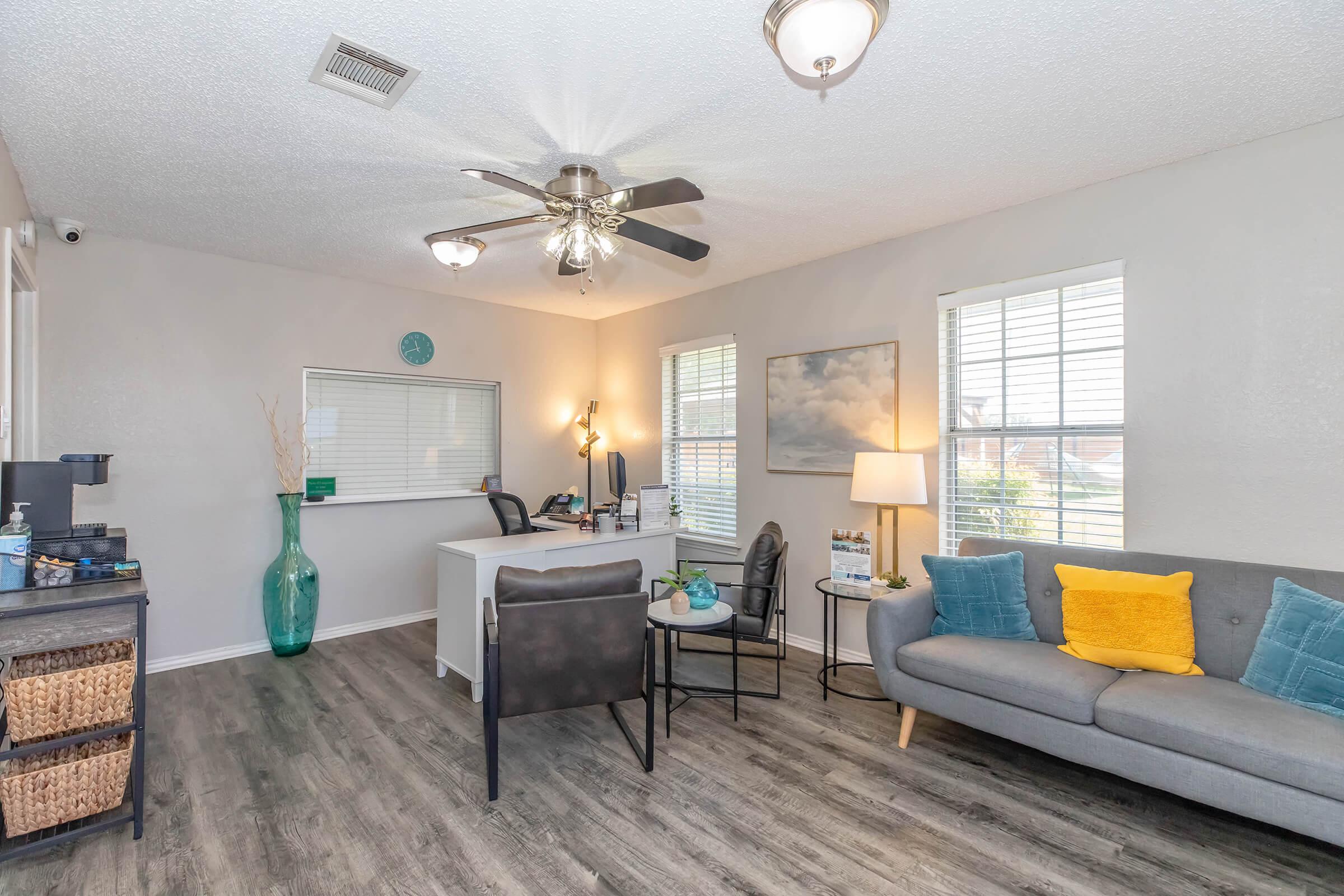 A cozy office space with a gray couch adorned with yellow and teal pillows, a small desk with a desktop computer, and a large plant in a glass vase. Natural light streams through two windows, adding warmth to the room, which features wooden flooring and a ceiling fan.