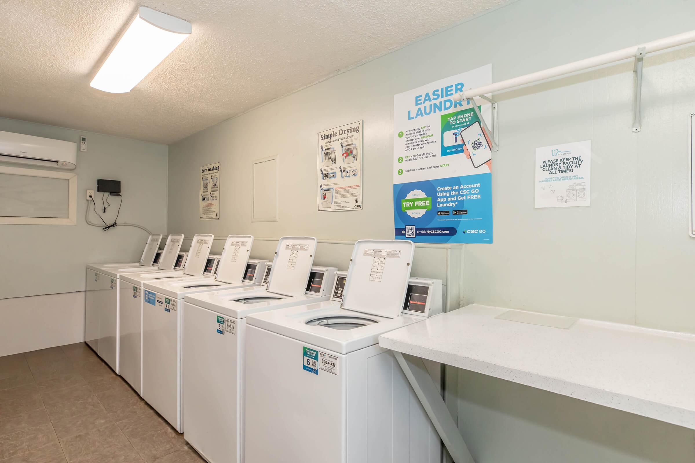 A clean and well-lit laundry room featuring several white washing machines, a folding counter, and posters with information about laundry services. The walls are painted a light color, and there is a ceiling light fixture providing illumination.