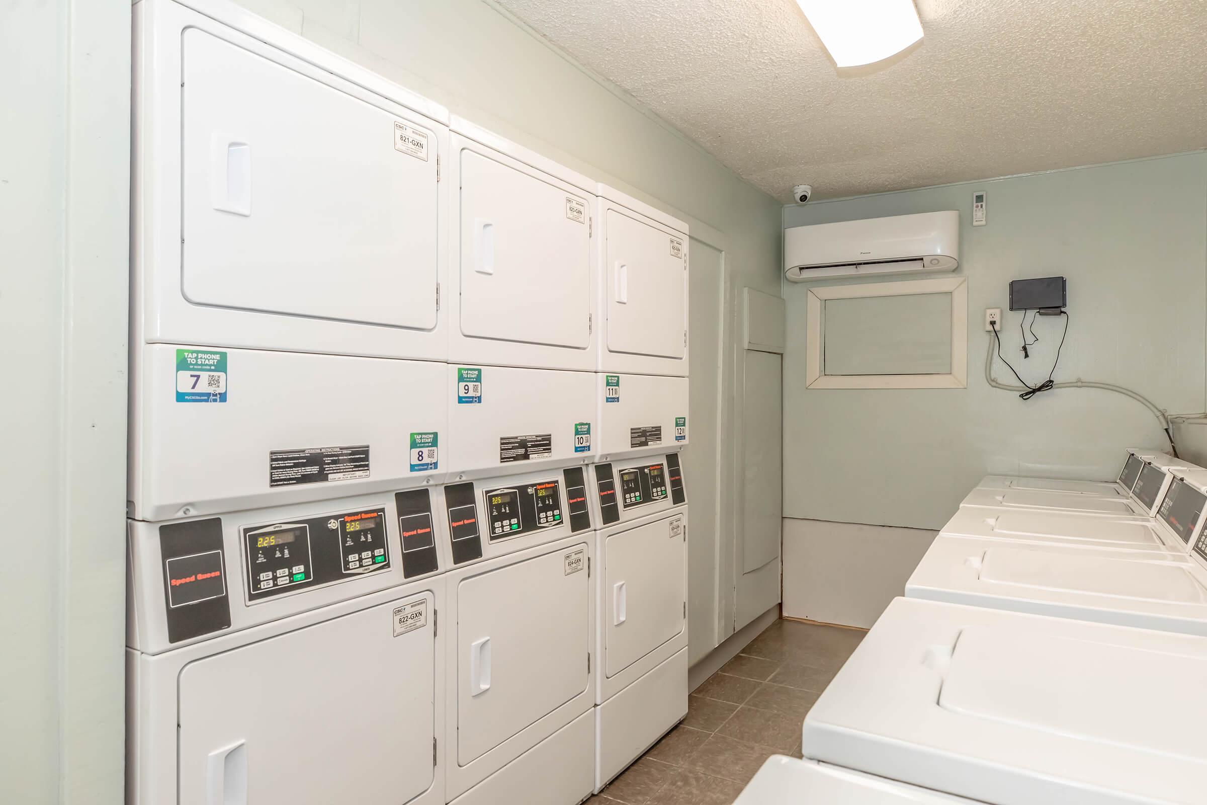 A clean, well-lit laundry room featuring several stacked washing machines and dryers against a pale green wall. The floor is tiled, and there is an air conditioning unit mounted on the wall. The space is organized and functional for laundry use.