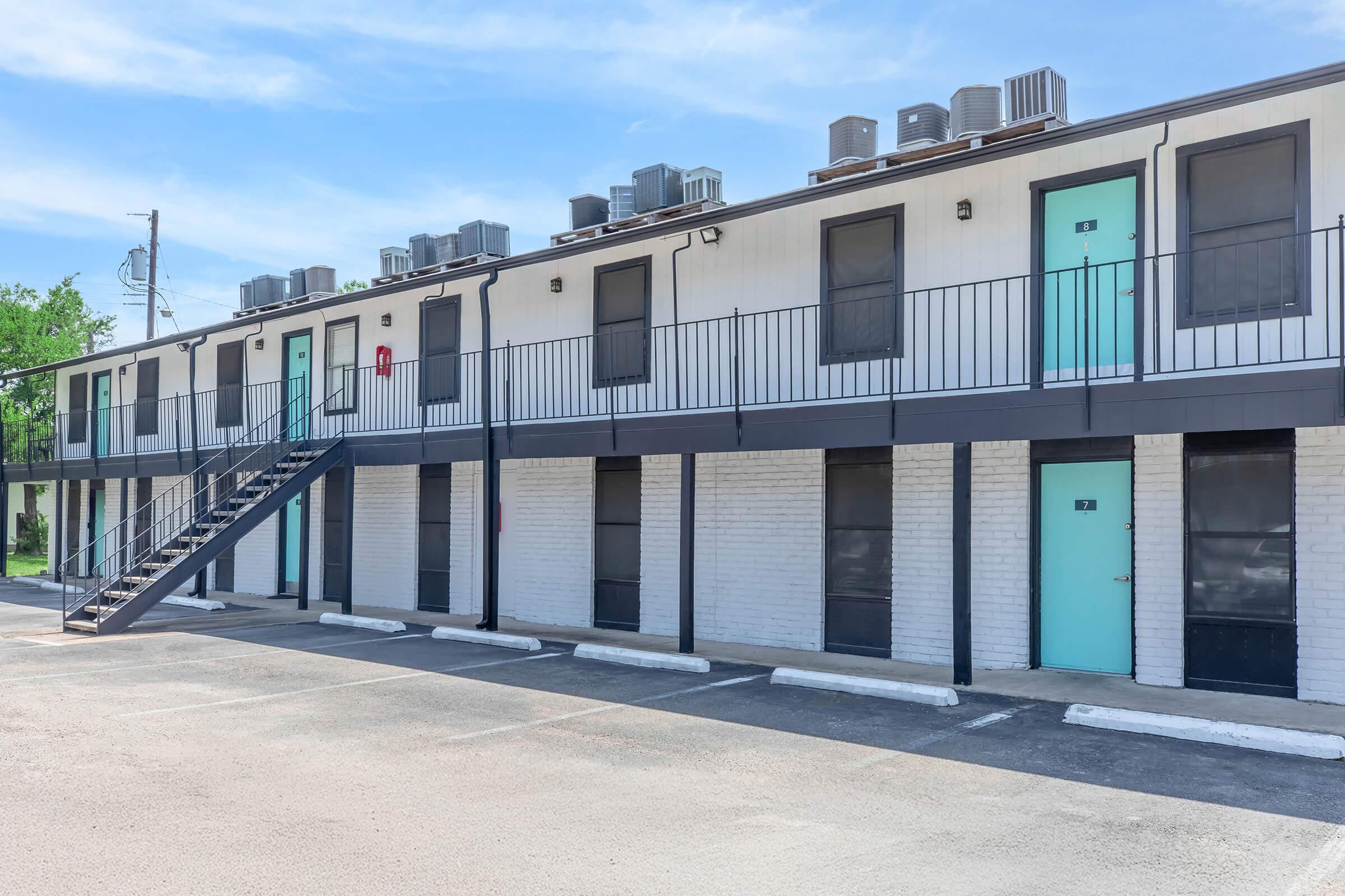 Two-story motel with a white brick exterior and black trim. Each unit features a turquoise door and has visible air conditioning units on the roof. A metal staircase leads to the second floor. Several parking spaces are available in front, and the sky is clear with a few clouds.