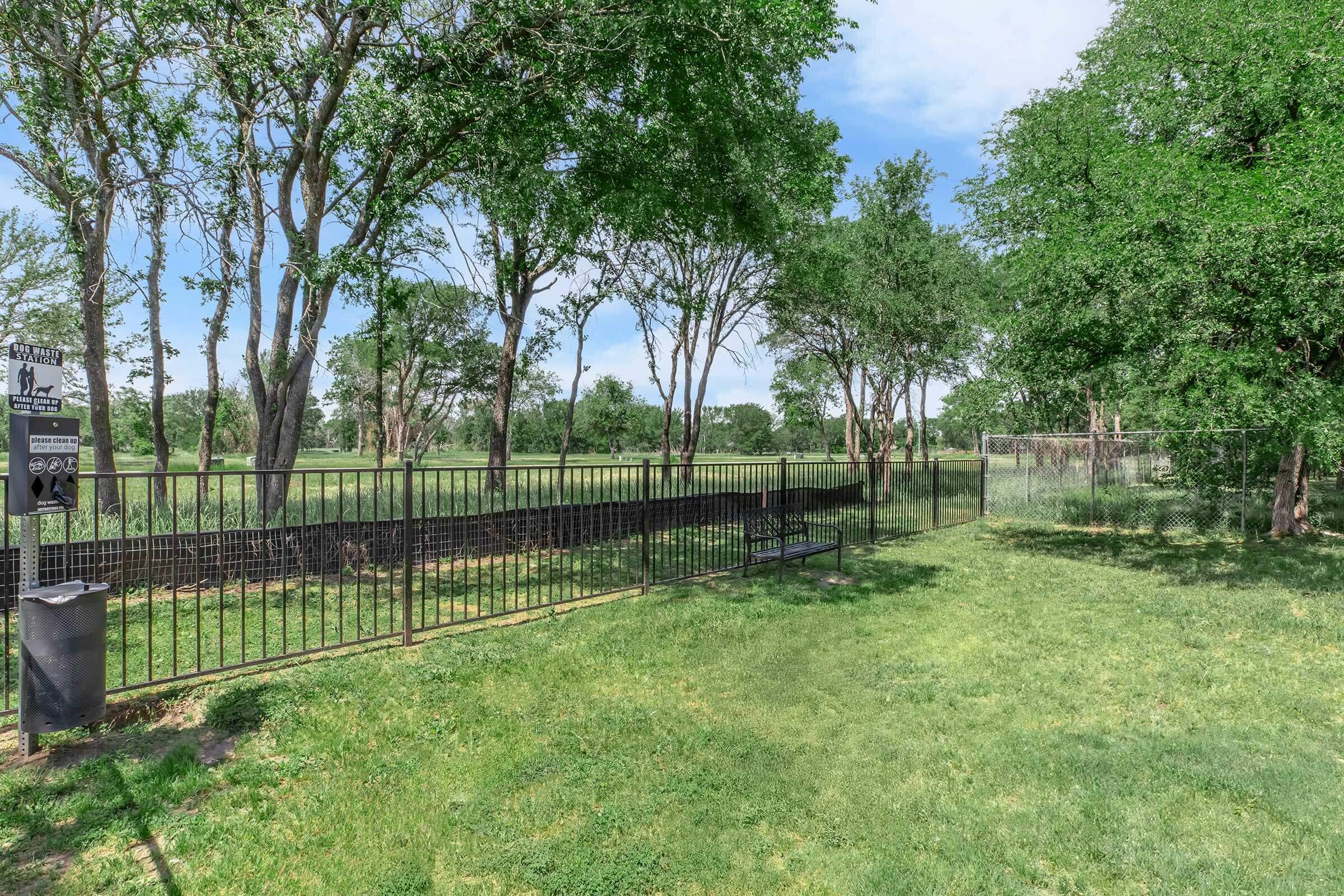 A green park area featuring a black metal fence bordering a grassy field. Several trees provide shade, and a bench is positioned along the path. In the background, a playground structure can be seen, indicating a family-friendly environment. The sky is clear with a few clouds.