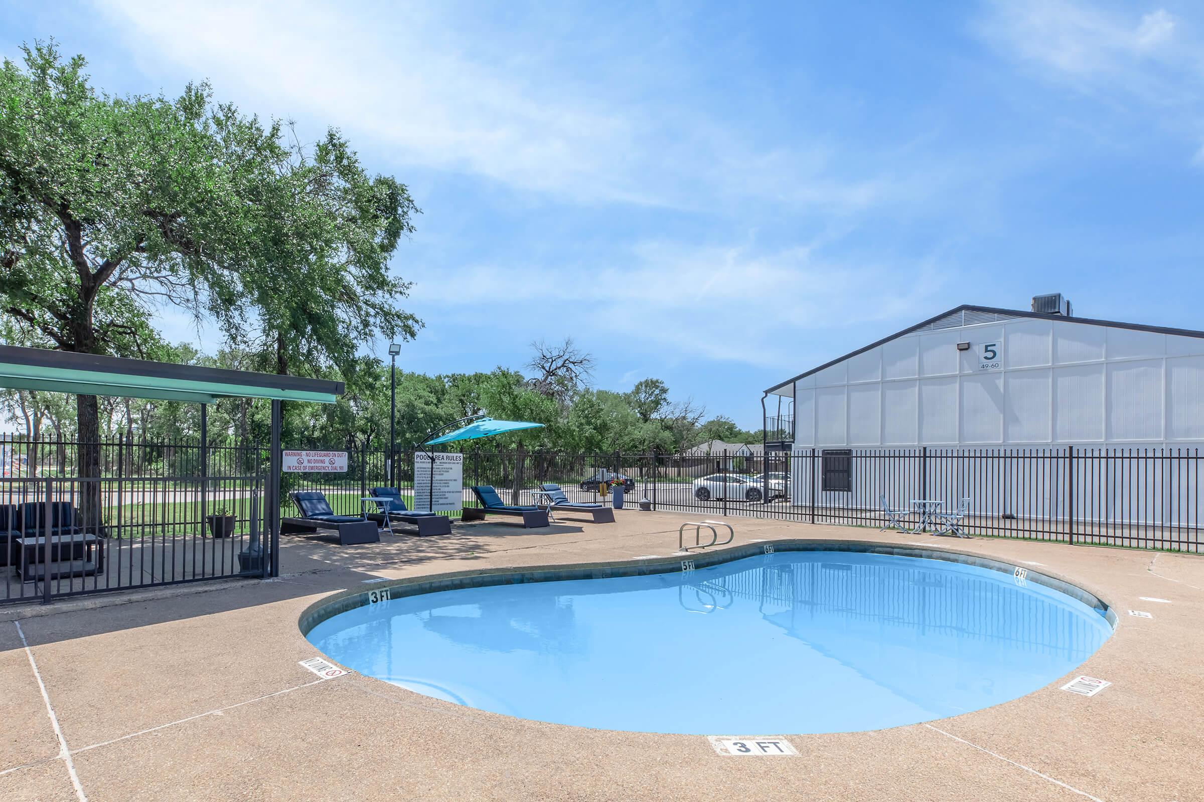 A clear blue swimming pool surrounded by lounge chairs and umbrellas. In the background, there is a fenced area with a building and trees. The sky is bright and sunny, creating a relaxing atmosphere.