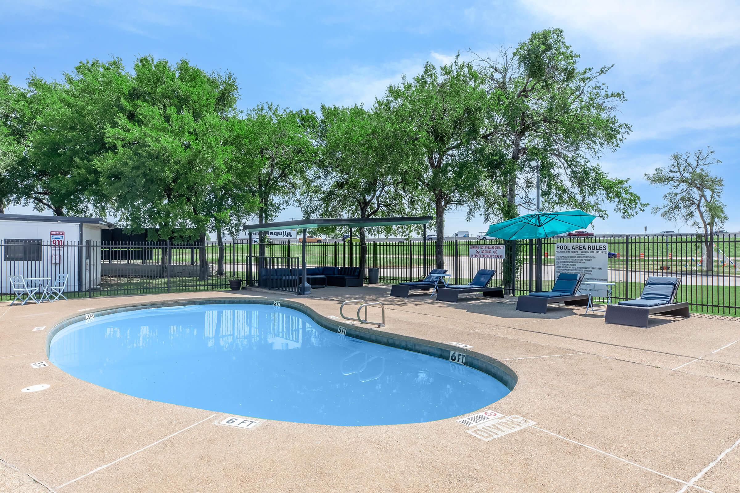 A clear swimming pool surrounded by lounge chairs and shaded by umbrellas, with greenery and a fenced area in the background. The pool area features a sign with pool rules, and the sky is blue with a few clouds. The scene is bright and inviting.