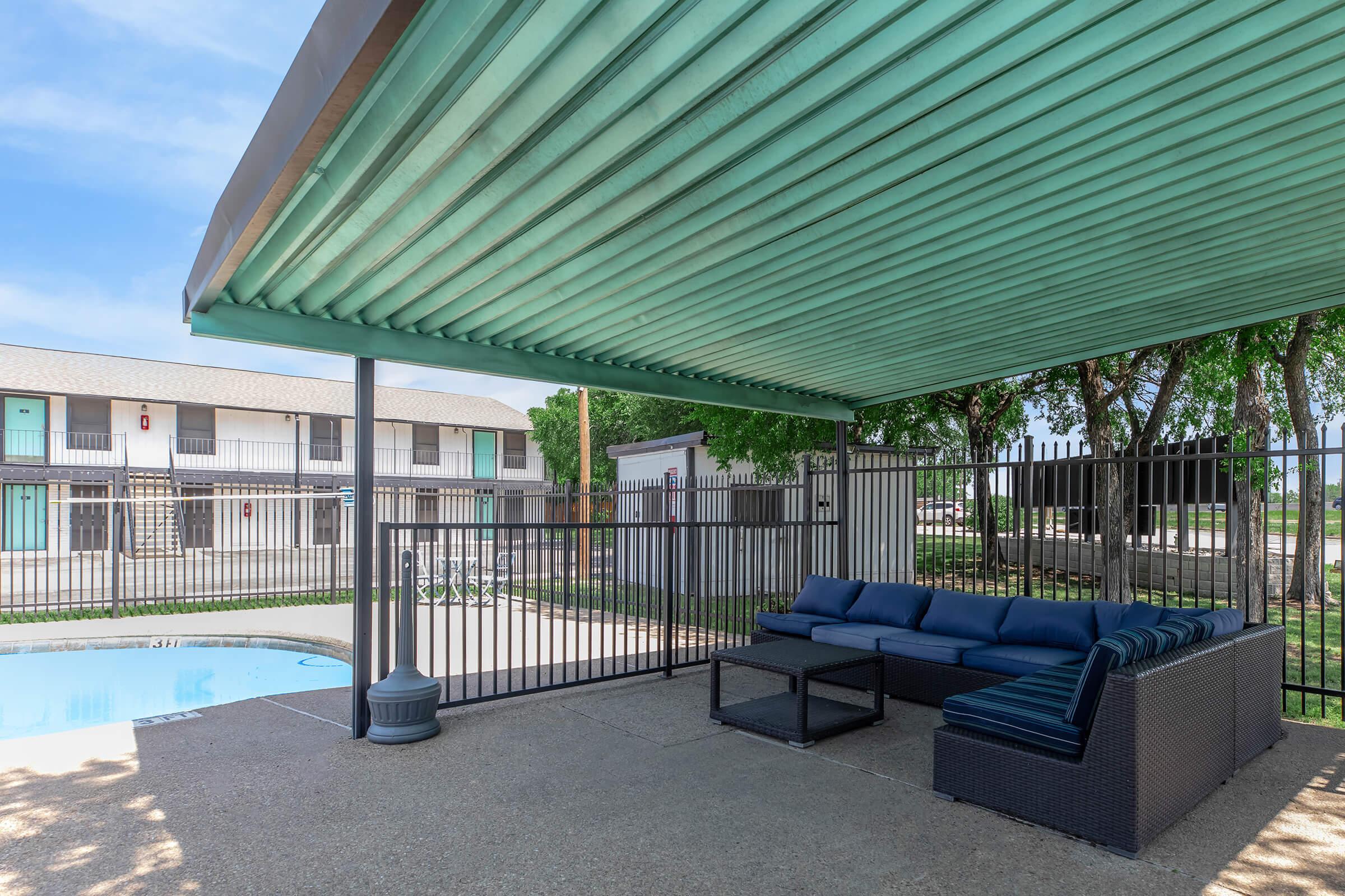 Covered outdoor seating area featuring a blue sofa set and a coffee table, adjacent to a swimming pool. The background includes a fenced pool area and a multi-story building with several rooms and green trees under a clear blue sky.