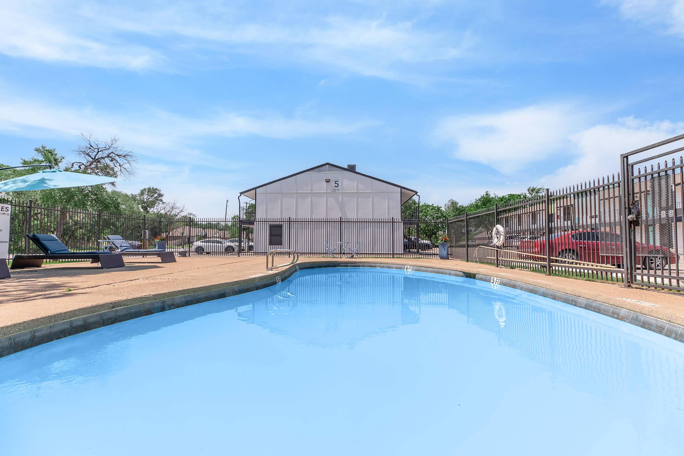 A clear blue swimming pool surrounded by a fence, with lounge chairs on one side. In the background, there is a modest building and trees lining the area under a bright, partly cloudy sky.