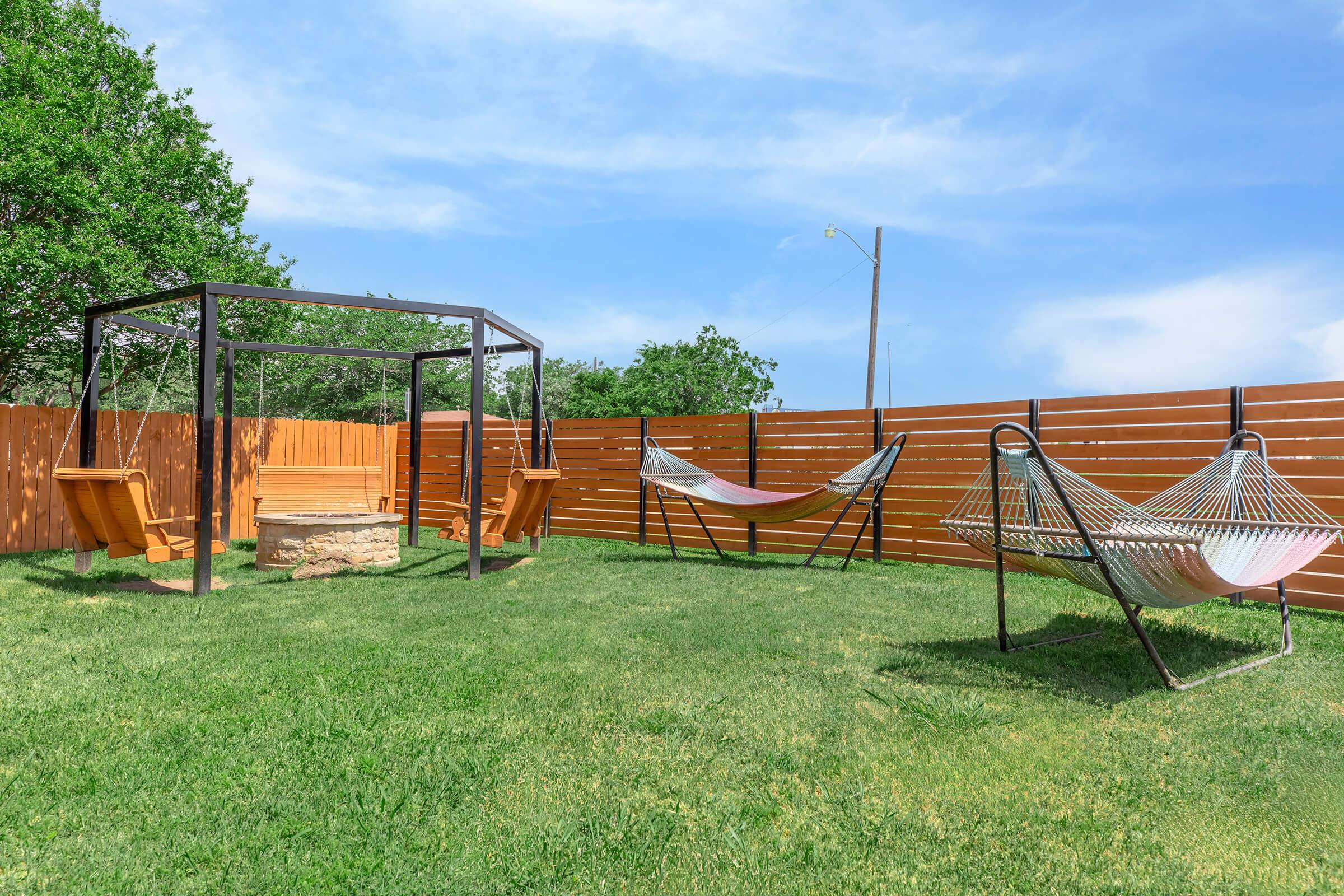 A backyard with a grassy area featuring a wooden swing set and two hammocks. The space is enclosed by a brown wooden fence, and there is a stone fire pit in the center. The sky is clear with a few clouds, and there are trees in the background, creating a peaceful outdoor setting.