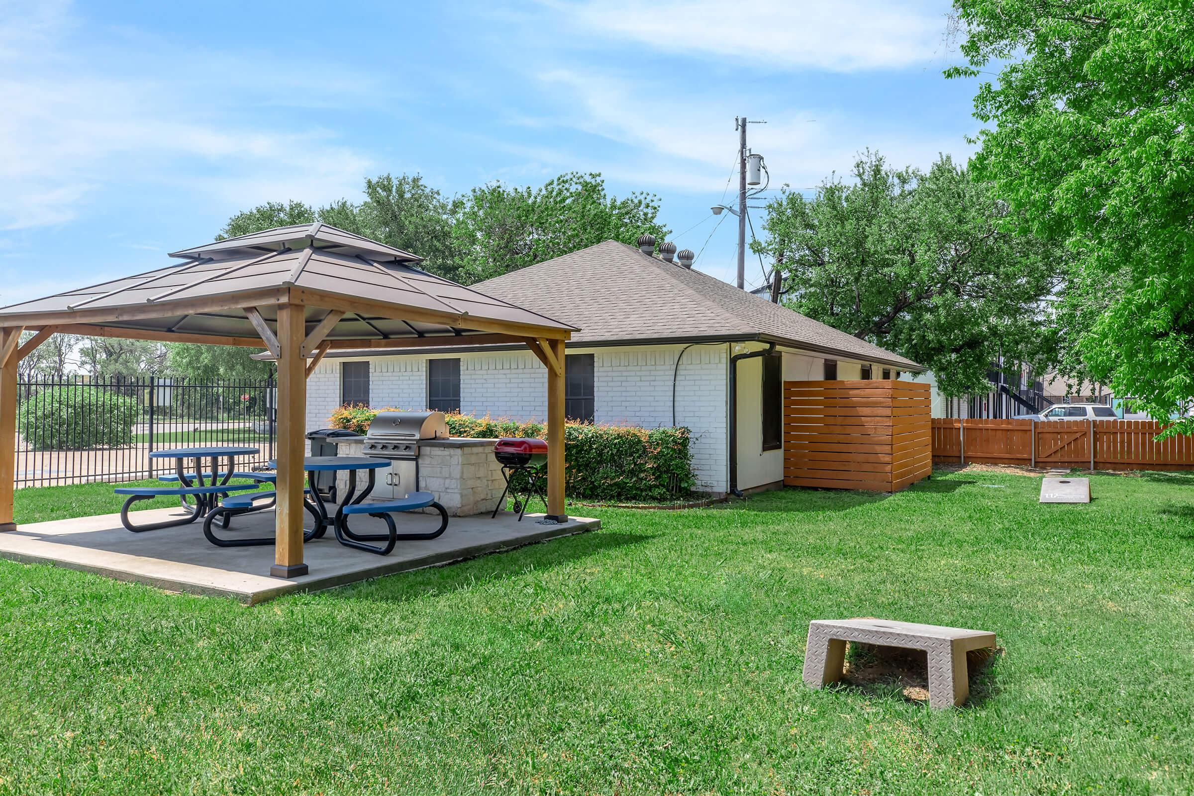 A spacious backyard featuring a gazebo with picnic tables, a grill, and well-maintained grass. In the background, there is a light-colored house and a wooden fence lining the property. Trees provide greenery, and a clear blue sky is visible above.