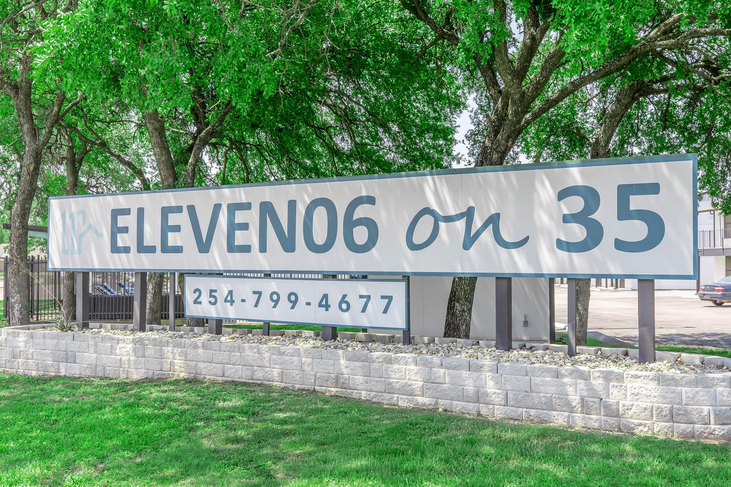 A large sign that reads "ELEVEN06 on 35" with a phone number "254-799-4677" below it. The sign is set against a backdrop of green grass and trees, indicating an outdoor location, likely a residential or commercial property.