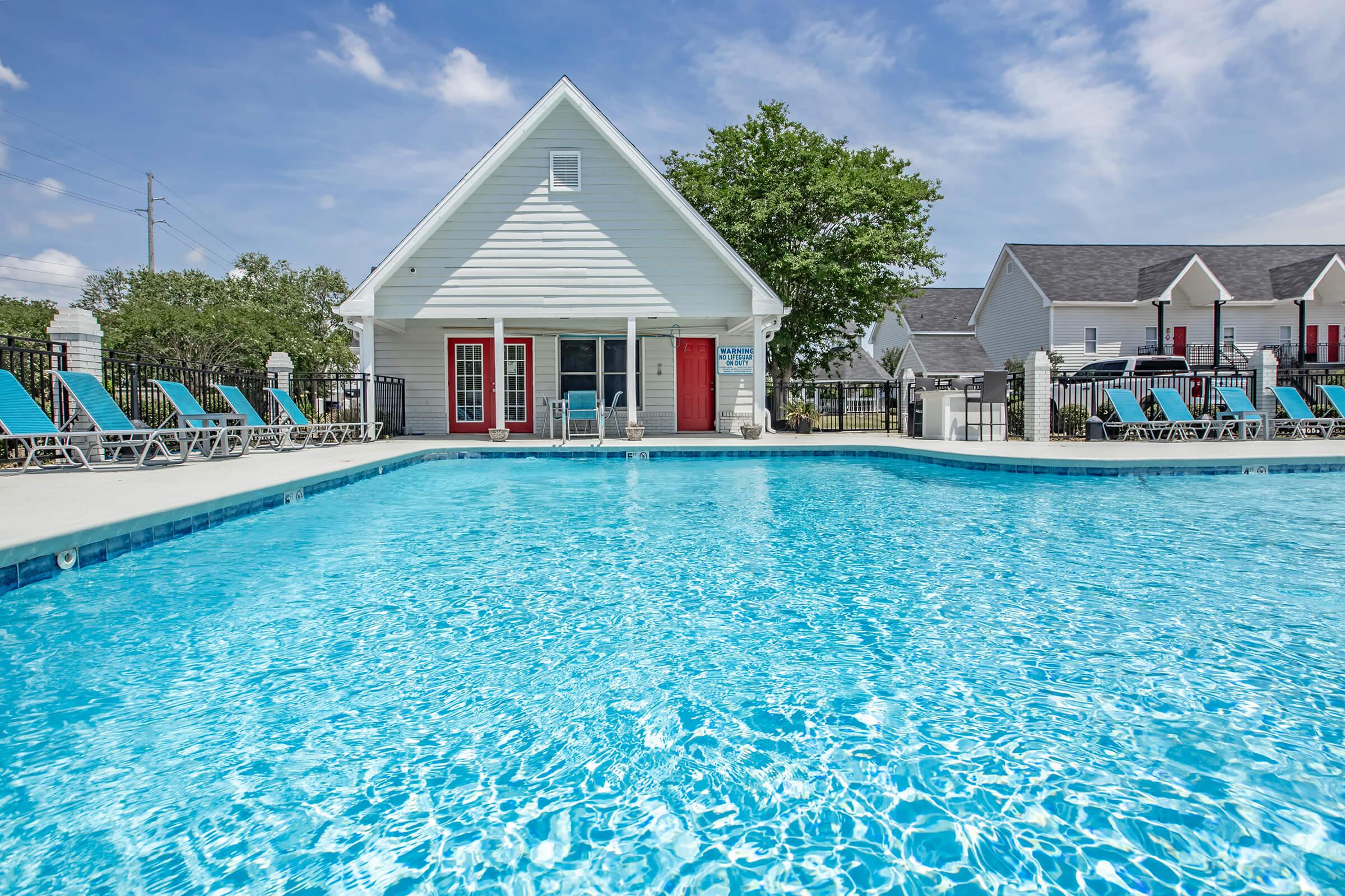 A clear, sparkling swimming pool with turquoise water in front of a white, two-story building featuring red doors. Surrounding the pool are multiple lounge chairs, and lush greenery can be seen in the background under a blue sky with scattered clouds. Perfect for leisure and relaxation.