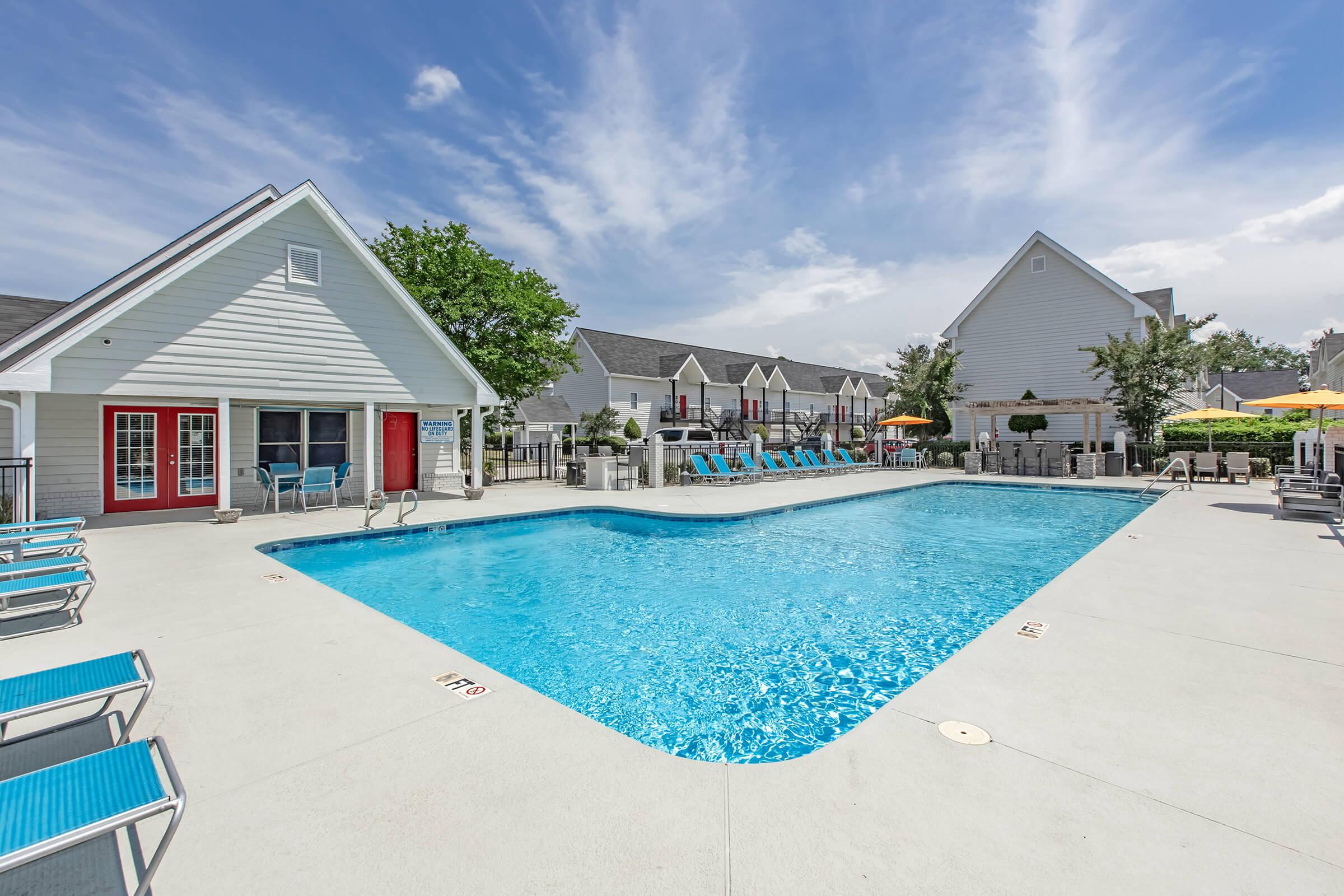 A bright outdoor pool area featuring a large, clear blue swimming pool surrounded by lounge chairs and umbrellas. White buildings with red doors are in the background, and trees add greenery to the setting. The sky is partly cloudy, creating a cheerful atmosphere.