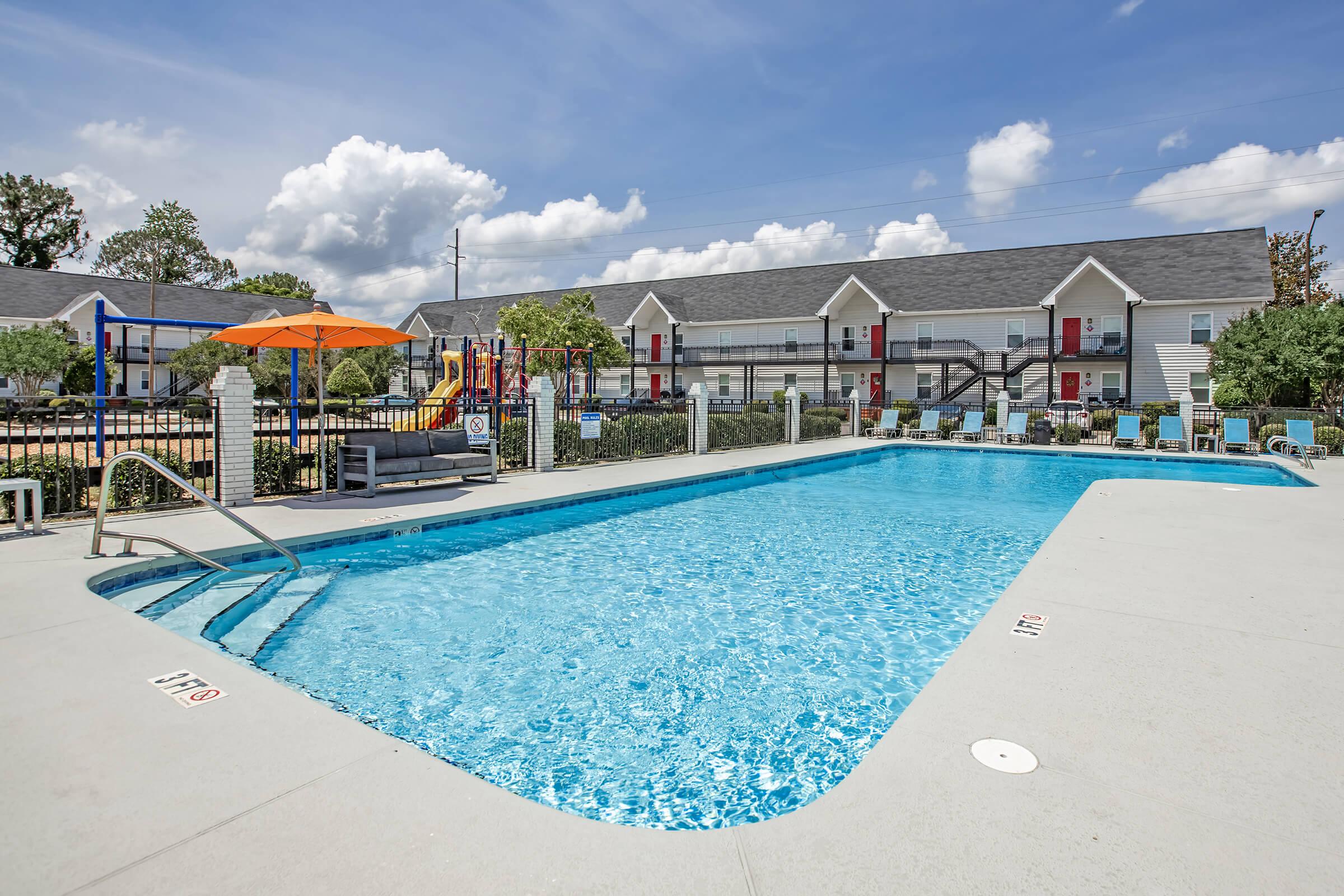 A vibrant swimming pool with clear blue water, surrounded by lounge chairs and an orange umbrella. In the background, there are two-story apartment buildings with red doors and lush greenery. The sky is bright with fluffy white clouds, creating a sunny, inviting atmosphere.
