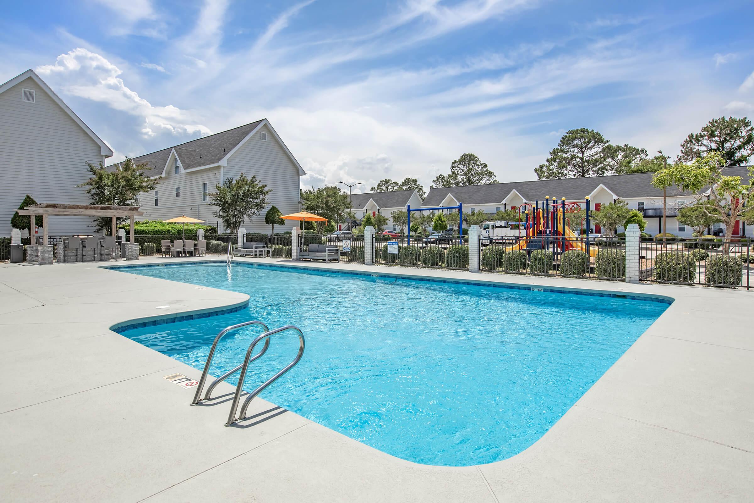 A clear blue swimming pool surrounded by a concrete deck, featuring two stair-handles. In the background, there are several residential buildings and a colorful playground visible. The sky is partly cloudy, and there are some trees and umbrellas providing shade around the area.