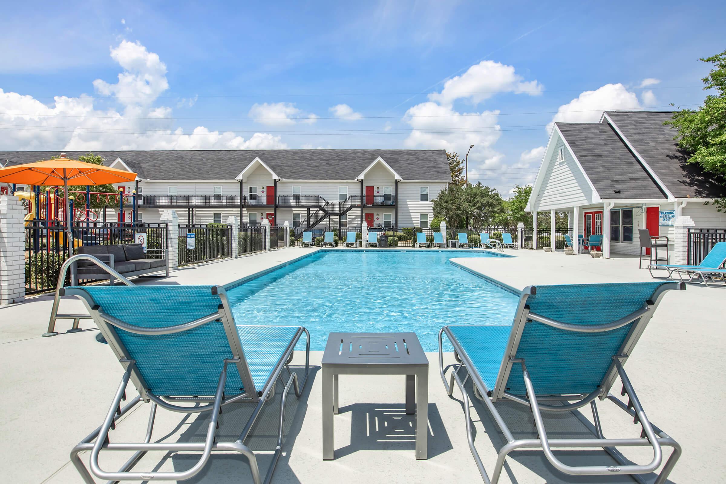 A bright outdoor swimming pool surrounded by lounge chairs, with an orange umbrella providing shade. The pool area is complemented by a well-maintained environment, and apartment buildings can be seen in the background against a clear blue sky with fluffy white clouds.