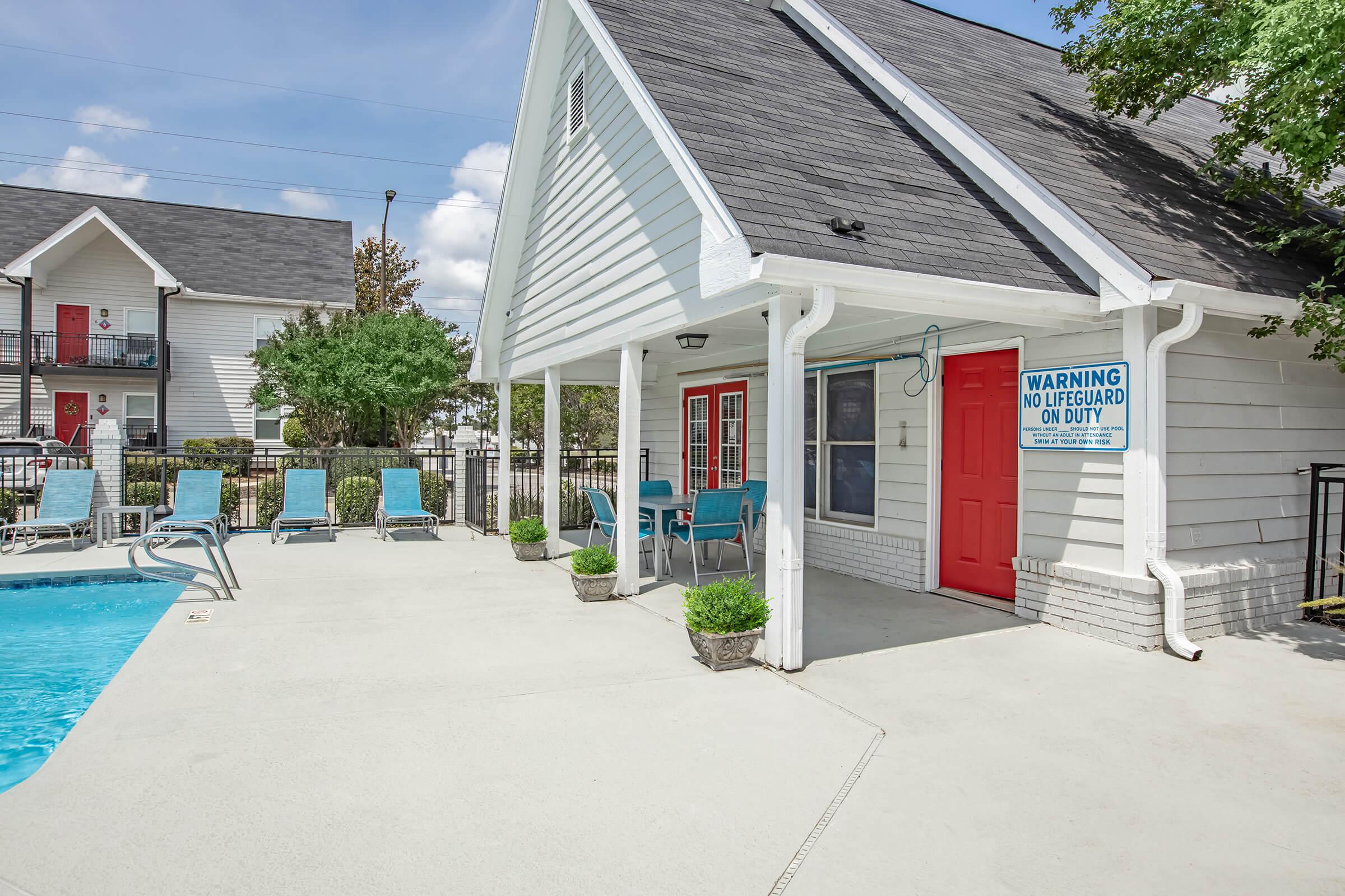 A well-lit pool area featuring a sparkling blue swimming pool, lounge chairs along the edge, and a nearby building with a red door. A sign warning "No Lifeguard on Duty" is visible. The scene is surrounded by greenery and a clear blue sky, creating a relaxing atmosphere.