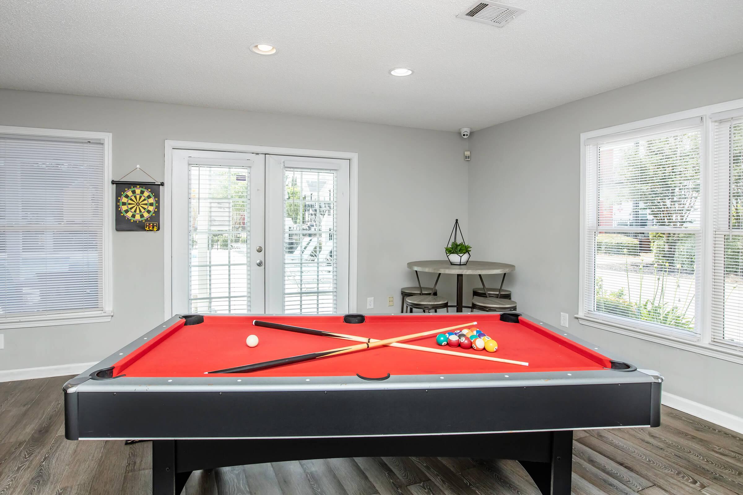 A bright, modern game room featuring a red pool table with a full set of balls and cues crossed on it. There are large windows letting in natural light, a dartboard on the wall, and a small seating area with two chairs near sliding glass doors leading outside. The room has neutral-colored walls and wooden flooring.