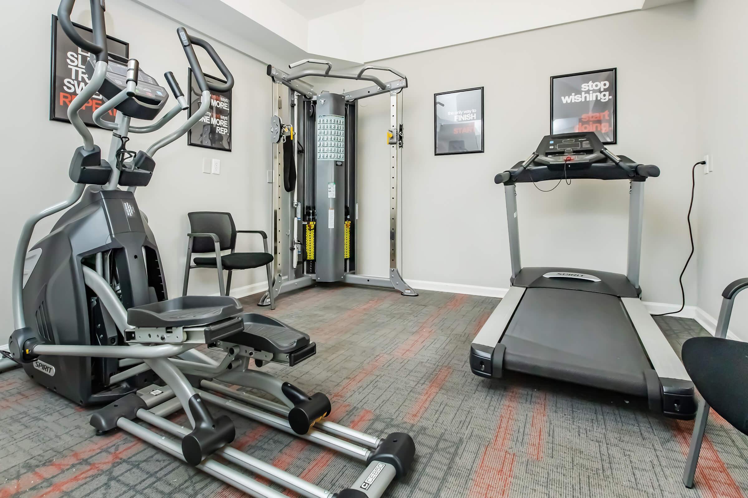 A fitness room featuring an exercise machine, a treadmill, and a black chair. The walls have motivational posters. The space is well-lit with carpeted flooring and modern decor, creating an inviting atmosphere for workouts.