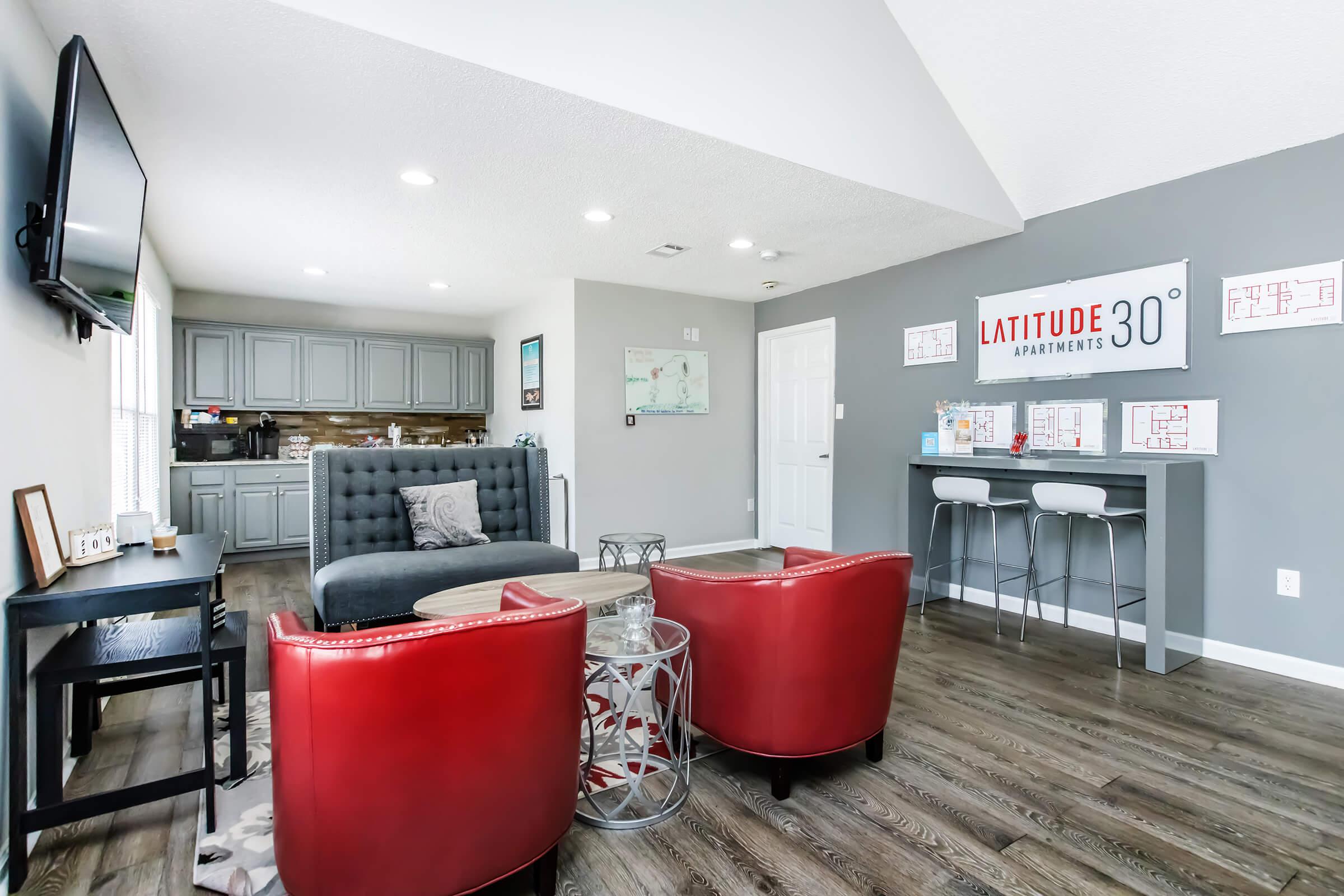 A modern apartment living space featuring a gray sofa, two red accent chairs, a coffee table, and a flat-screen TV mounted on the wall. The kitchen area is visible, showcasing gray cabinets and a stylish backsplash. A sign for "Latitude 30 Apartments" is displayed on the wall. The overall decor is contemporary and inviting.