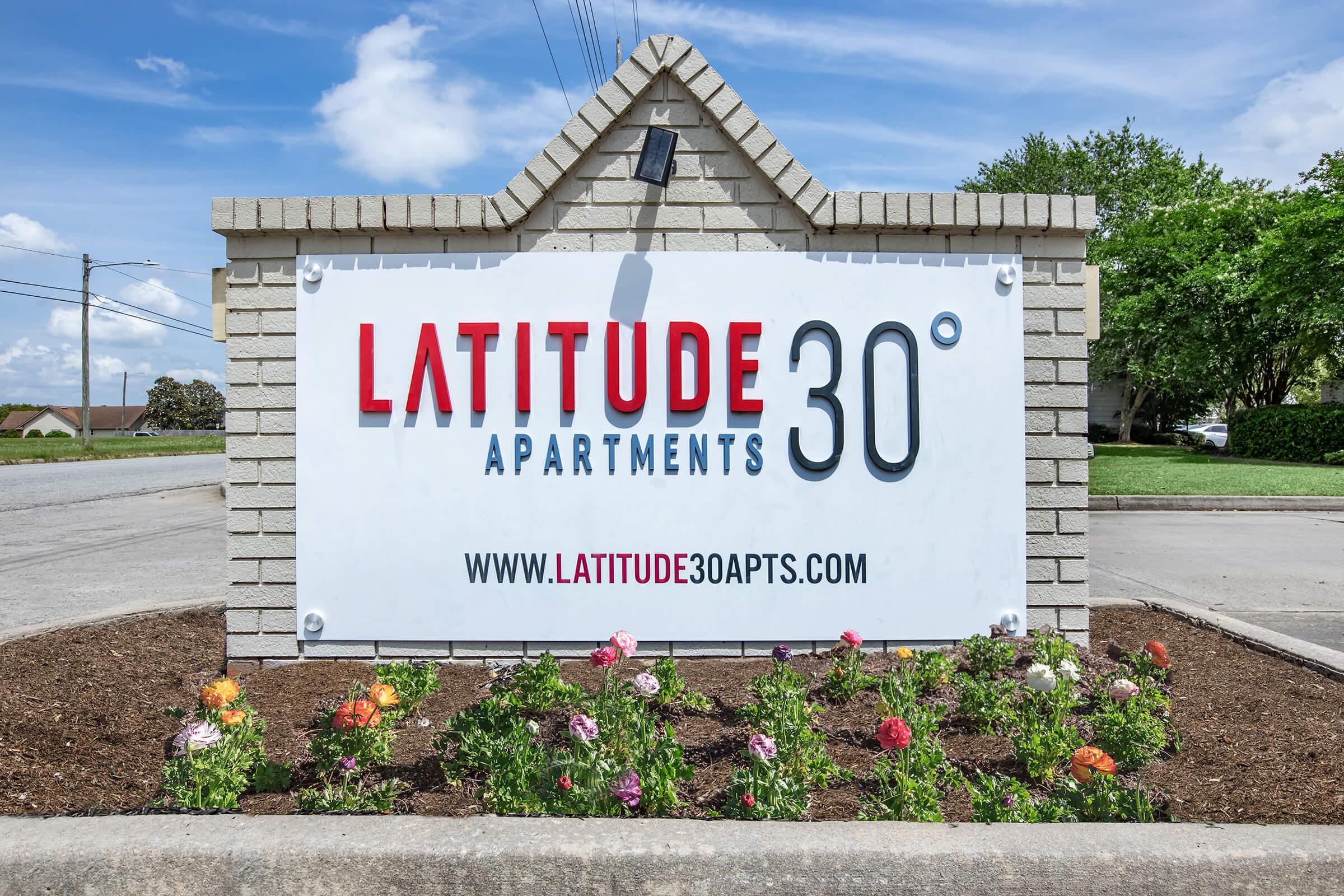 Sign for Latitude 30° Apartments featuring the name prominently displayed in bold letters. The sign includes a web address at the bottom and is surrounded by vibrant flowers and greenery, set against a backdrop of a clear blue sky.