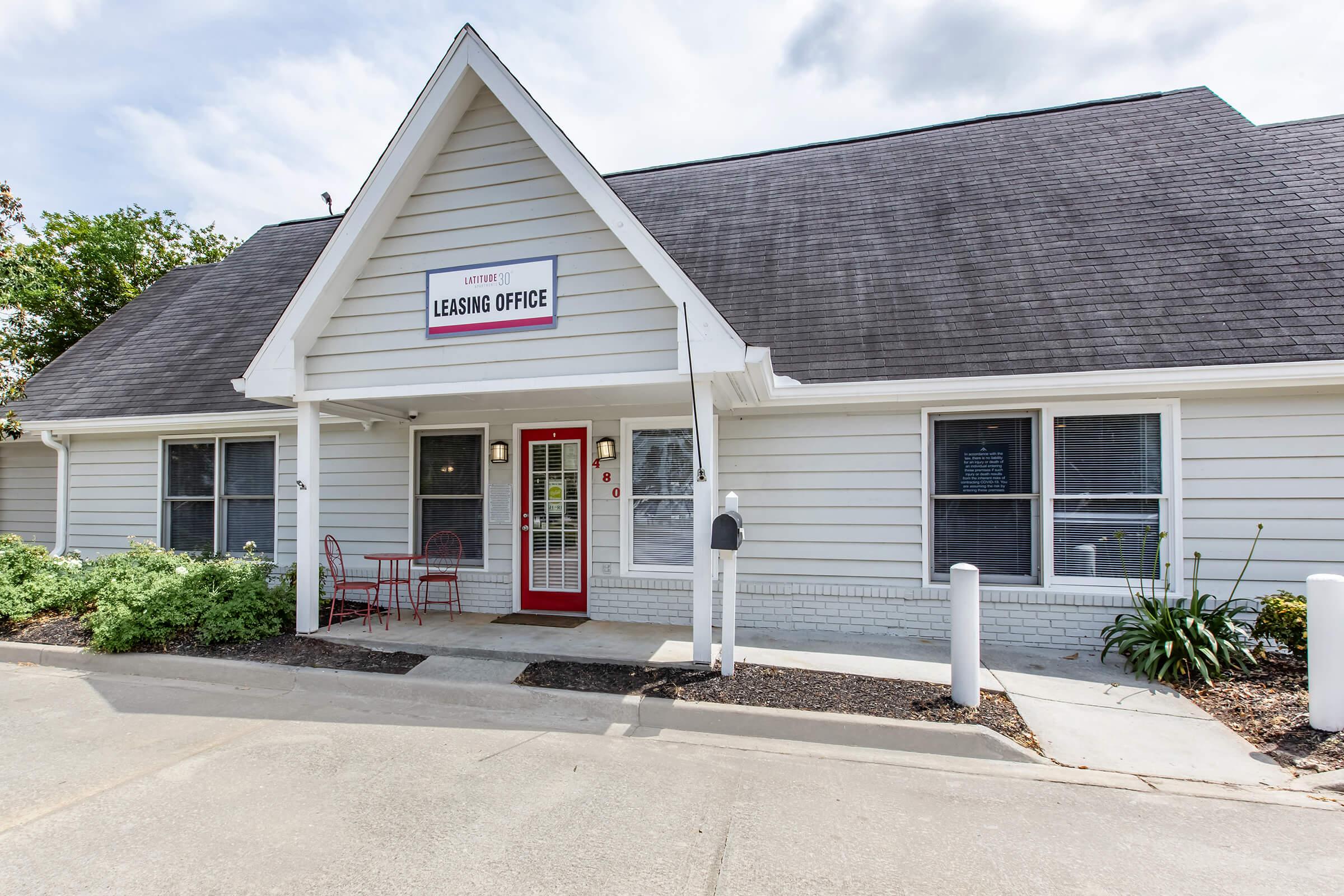 A modern leasing office with a triangular roof and white siding. The building features a red door, a sign labeled "Leasing Office," and a small patio with two chairs out front. Neatly trimmed landscaping surrounds the entrance, and a mailbox is visible nearby.