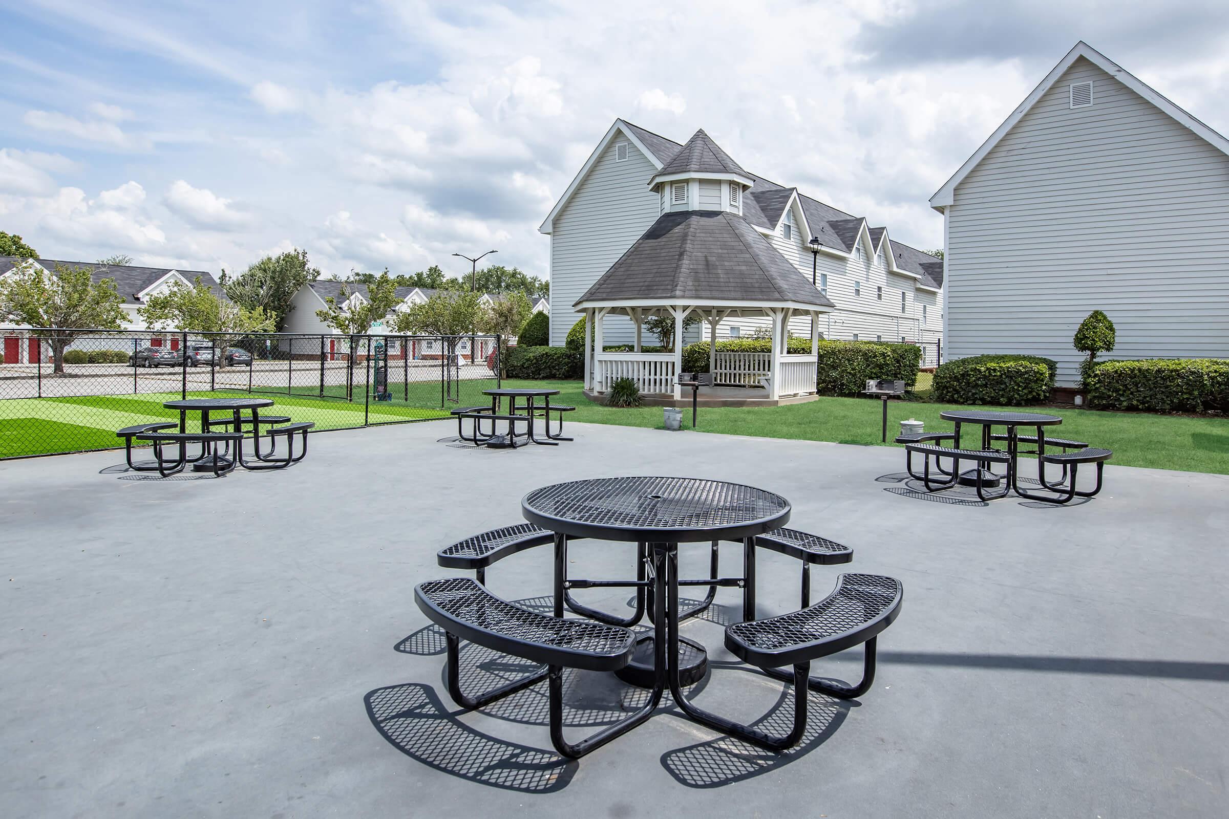 Outdoor recreational area featuring multiple black metal picnic tables on a paved surface. In the background, a white gazebo is visible, along with residential houses and neatly trimmed grass. The sky is partly cloudy, creating a pleasant atmosphere for social gatherings.