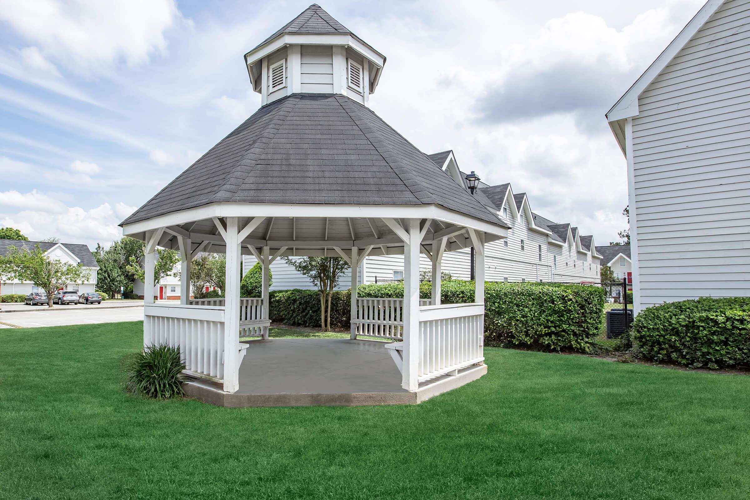 A charming white gazebo with a gray roof, situated in a green lawn area. Surrounding the gazebo are neatly trimmed hedges and a row of residential buildings in the background under a partly cloudy sky. The scene conveys a serene outdoor space suitable for relaxation.